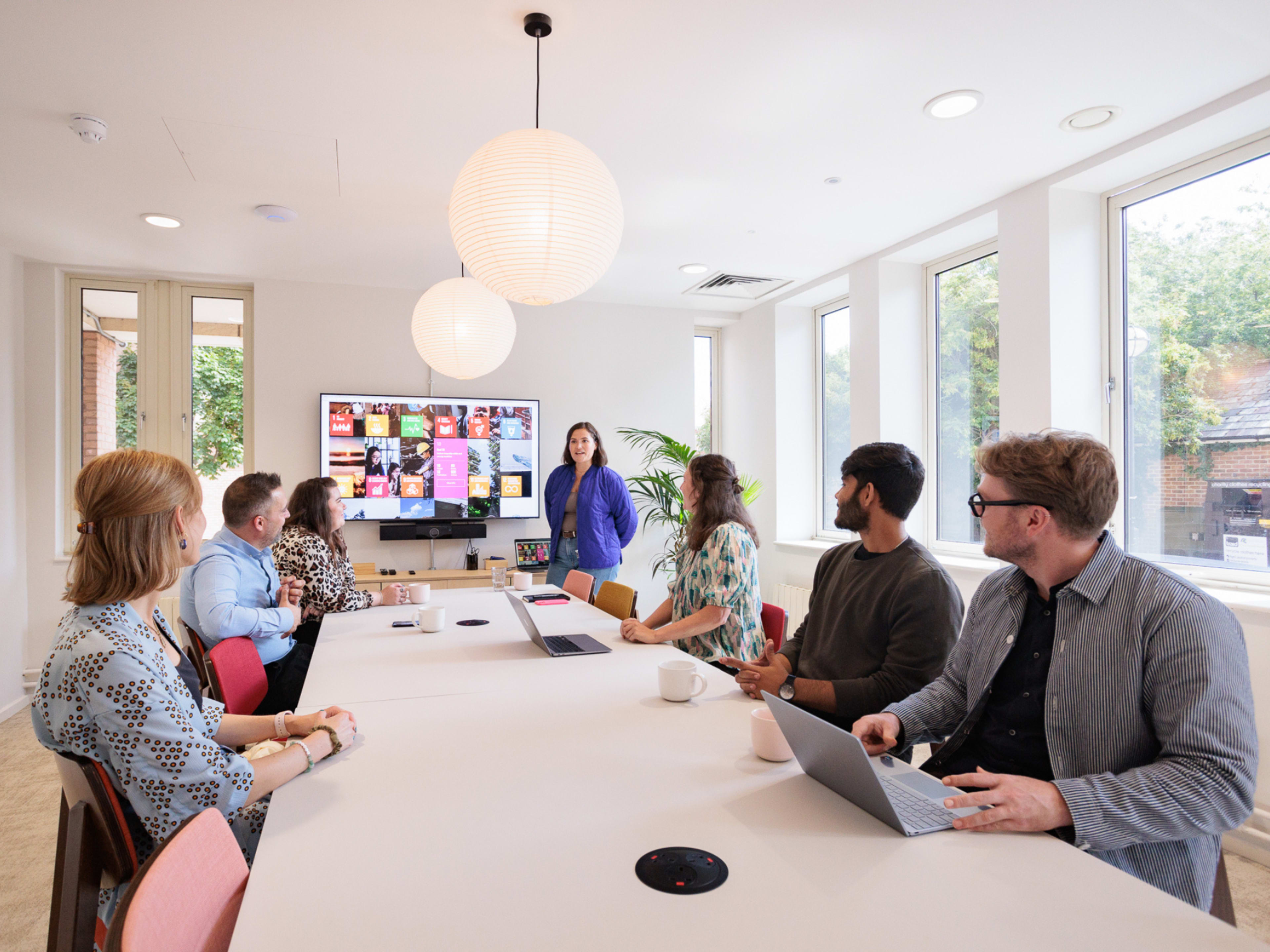 A business meeting is taking place in a brightly lit room, where a woman is presenting to a group seated around a table, with a large screen displaying various visuals behind her.