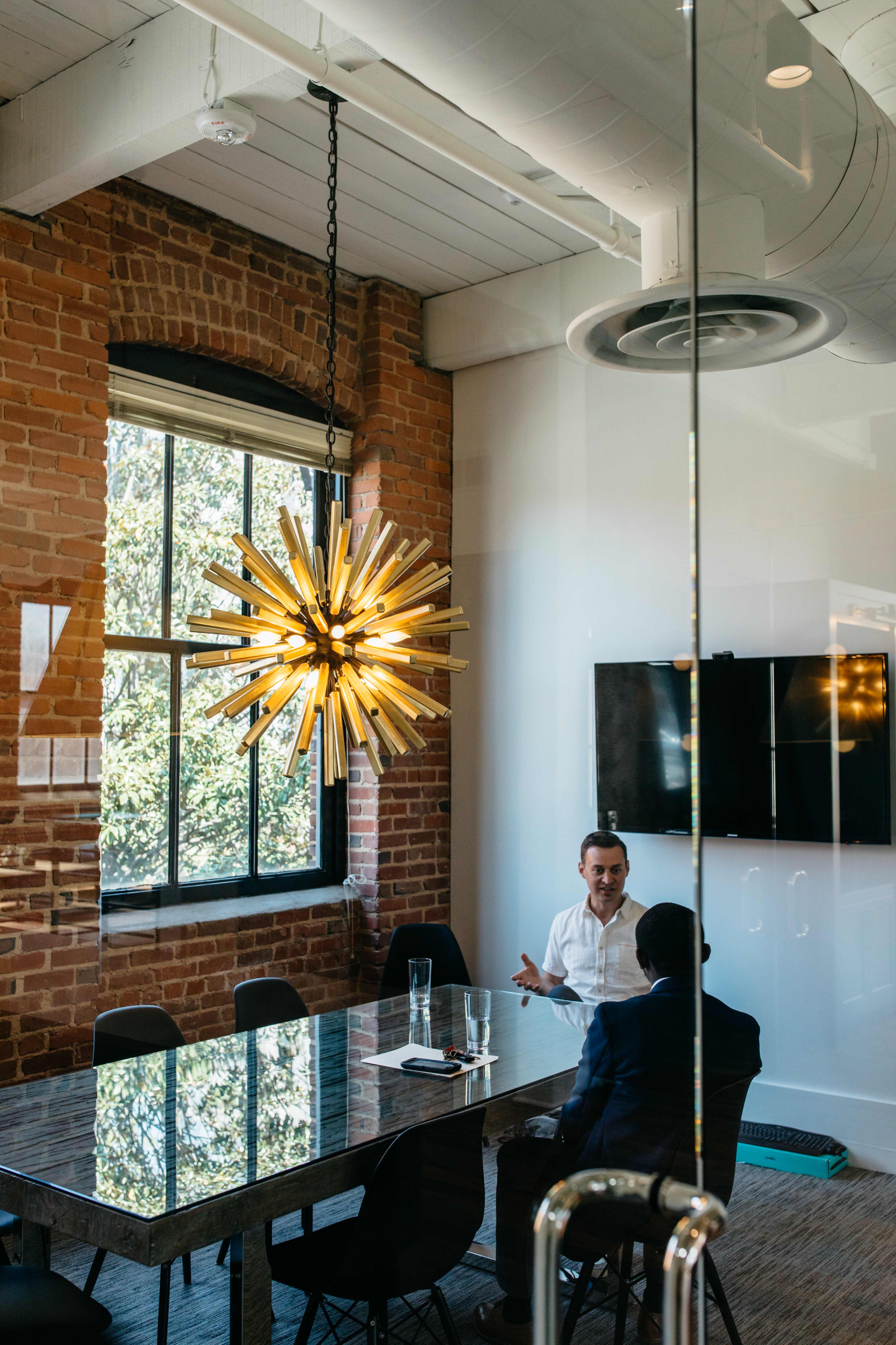 A glass-walled meeting room features a wooden table, two men engaged in conversation, and a large, starburst chandelier above.