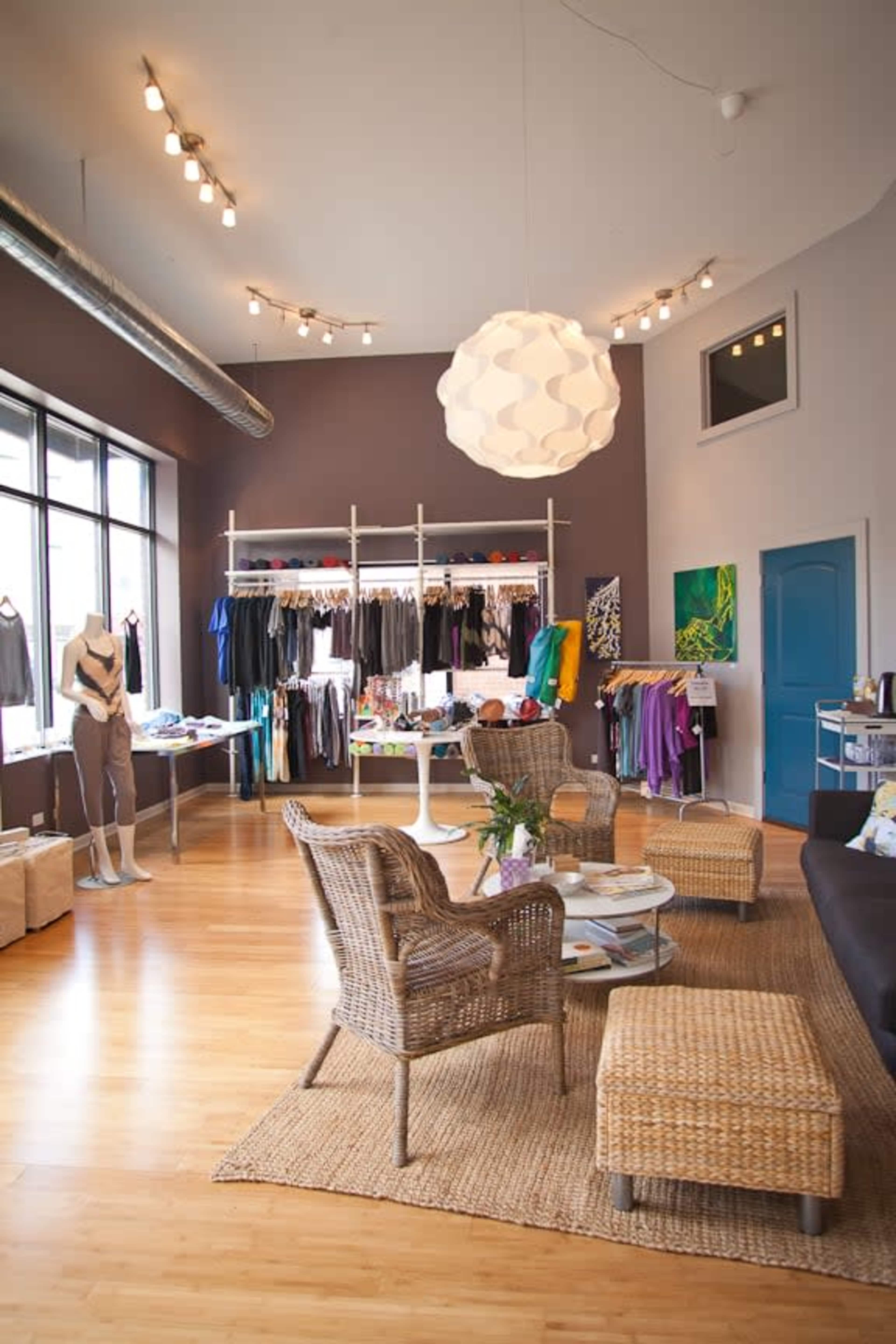 The image shows a modern retail space featuring clothing racks, a seating area with wicker furniture, and a large light fixture overhead.