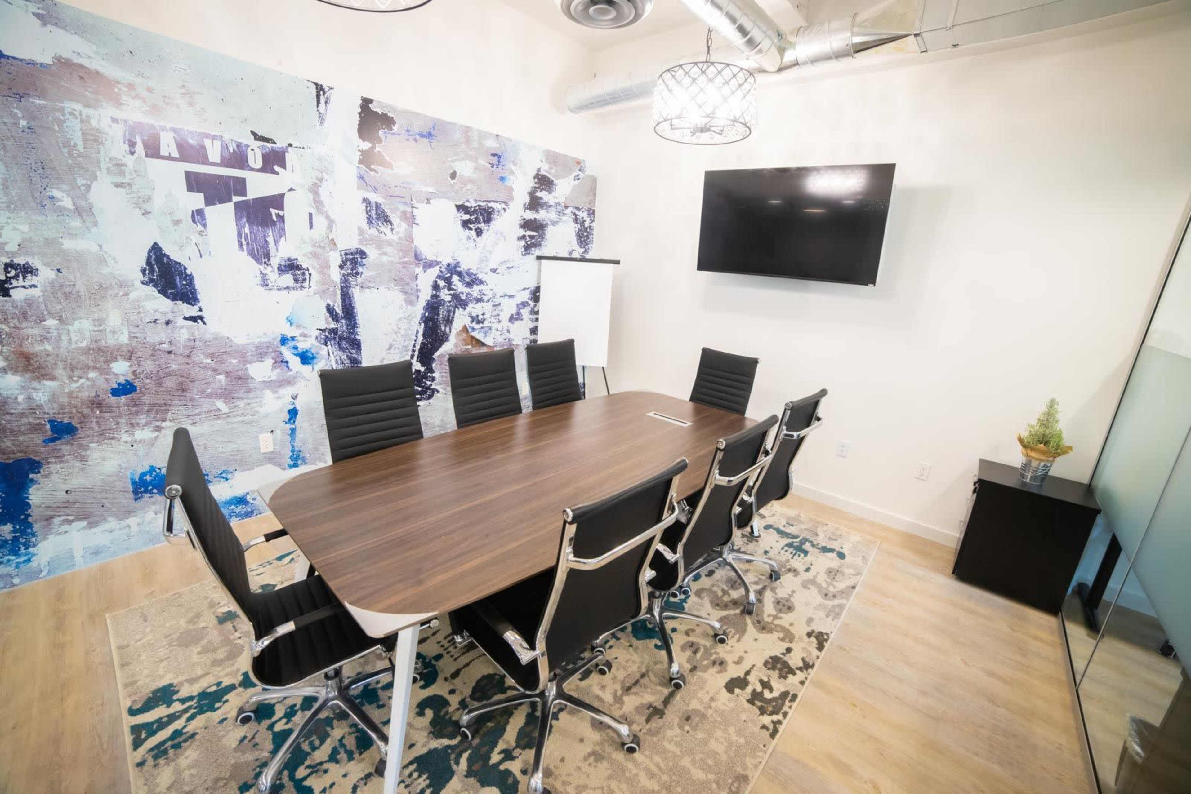 The image shows a modern conference room with a large wooden table surrounded by black chairs, a wall featuring abstract artwork, and a television mounted on the wall.