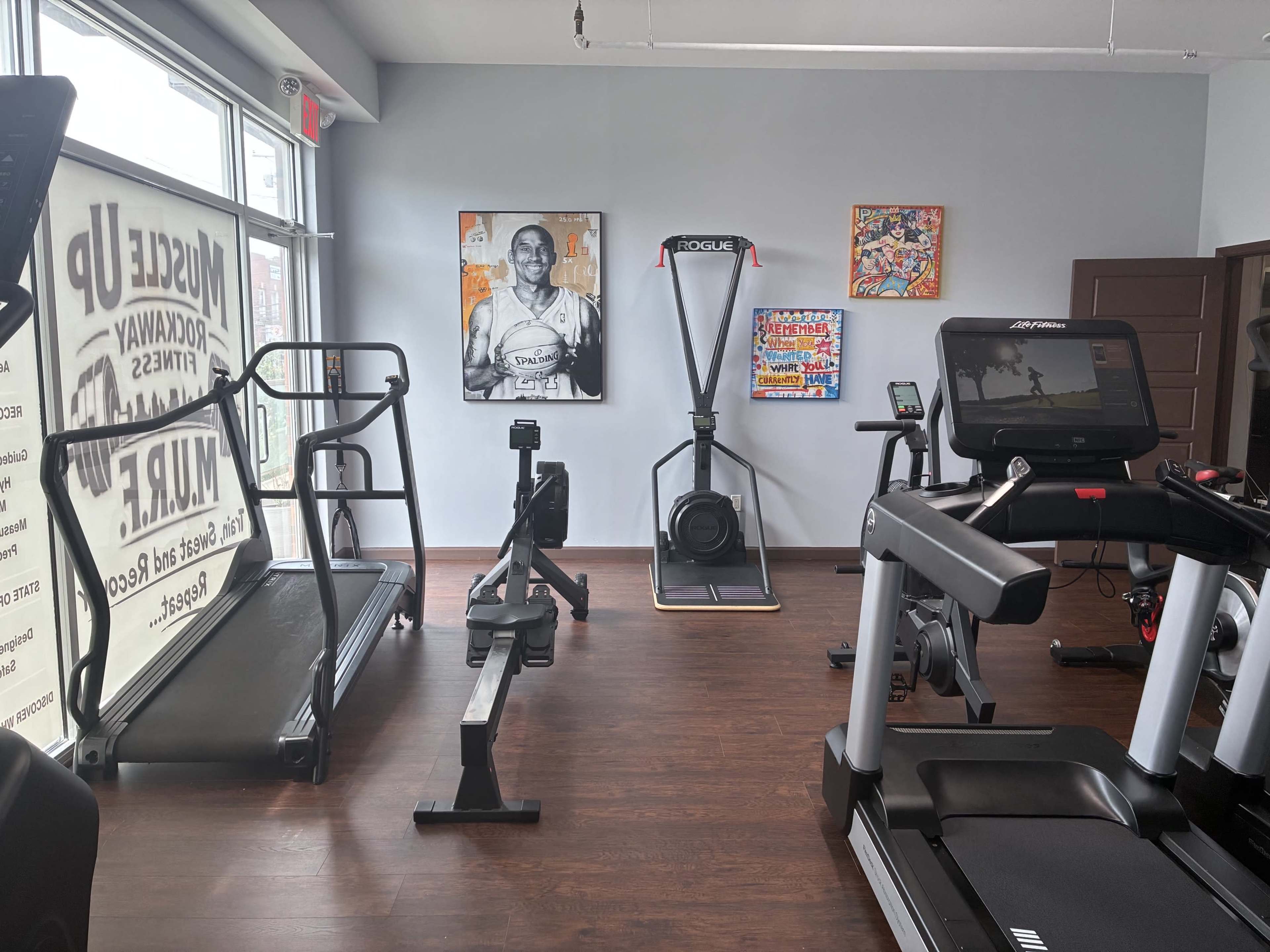 The image shows a fitness studio equipped with various exercise machines, including treadmills, an elliptical, and a rowing machine, along with artwork on the walls.