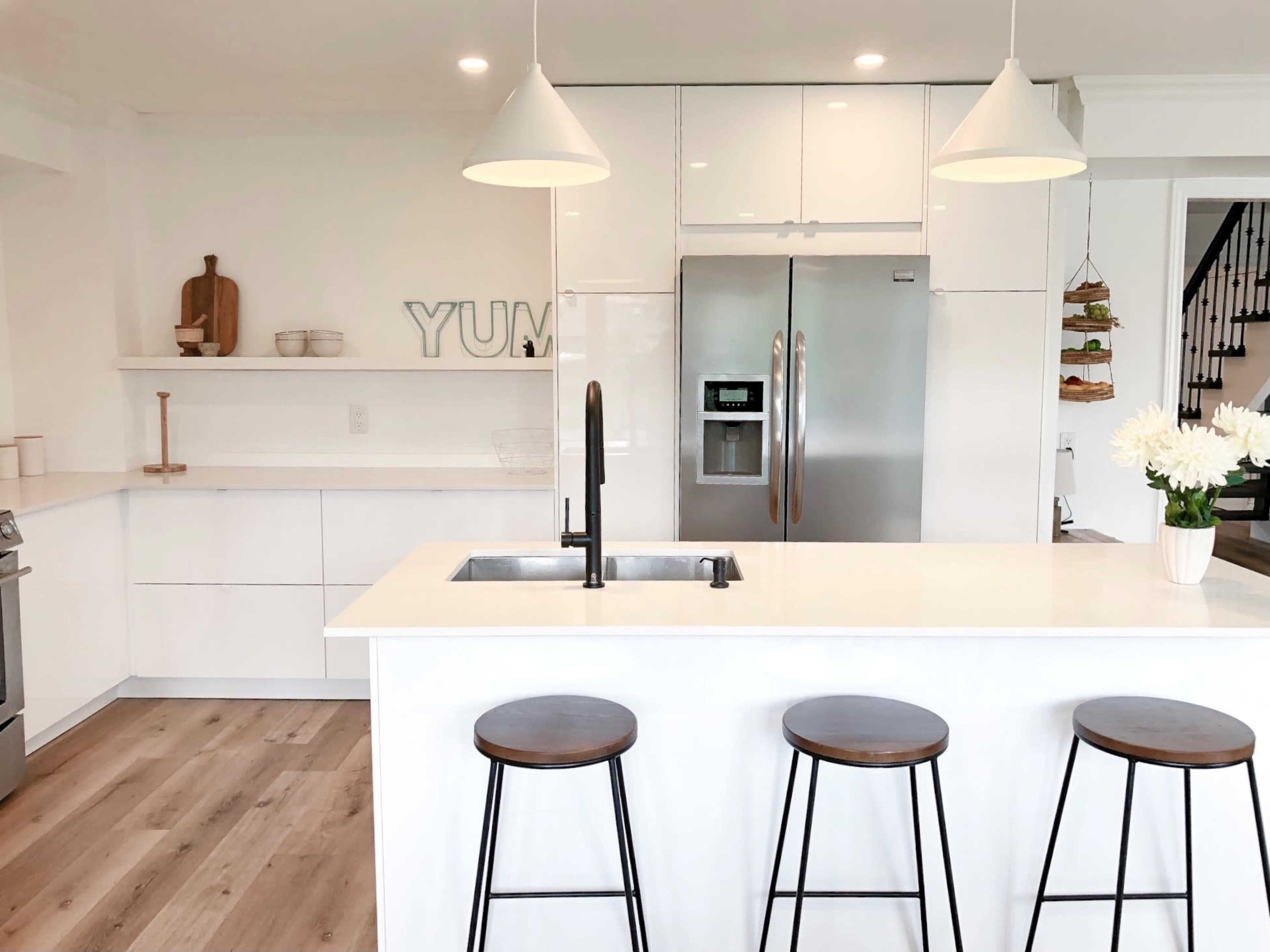 A modern kitchen with a white island, three wooden stools, stainless steel appliances, and minimalistic decor.