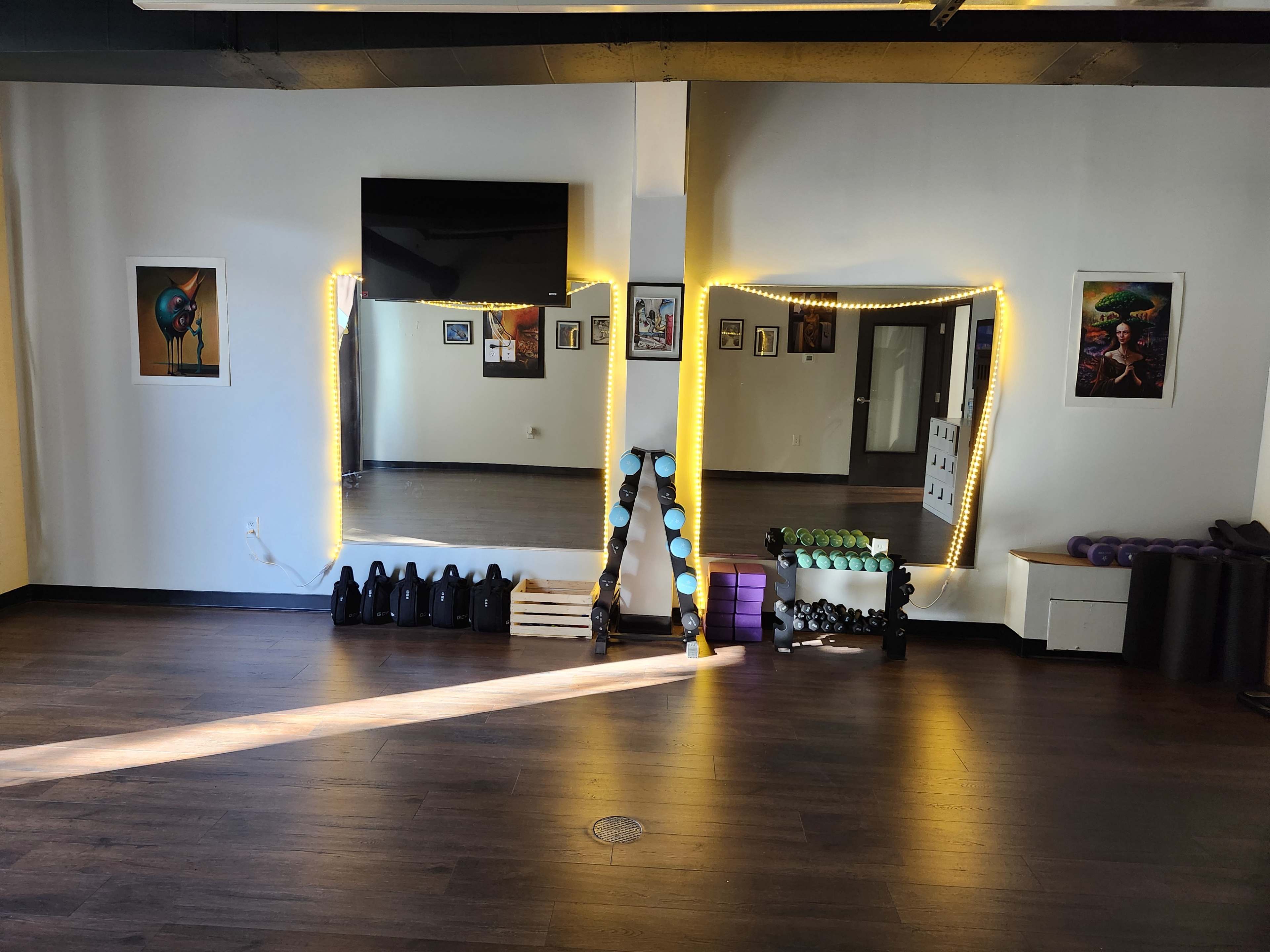 The image shows a fitness studio with a mirrored wall, exercise equipment arranged on the floor, and artwork displayed on the walls.