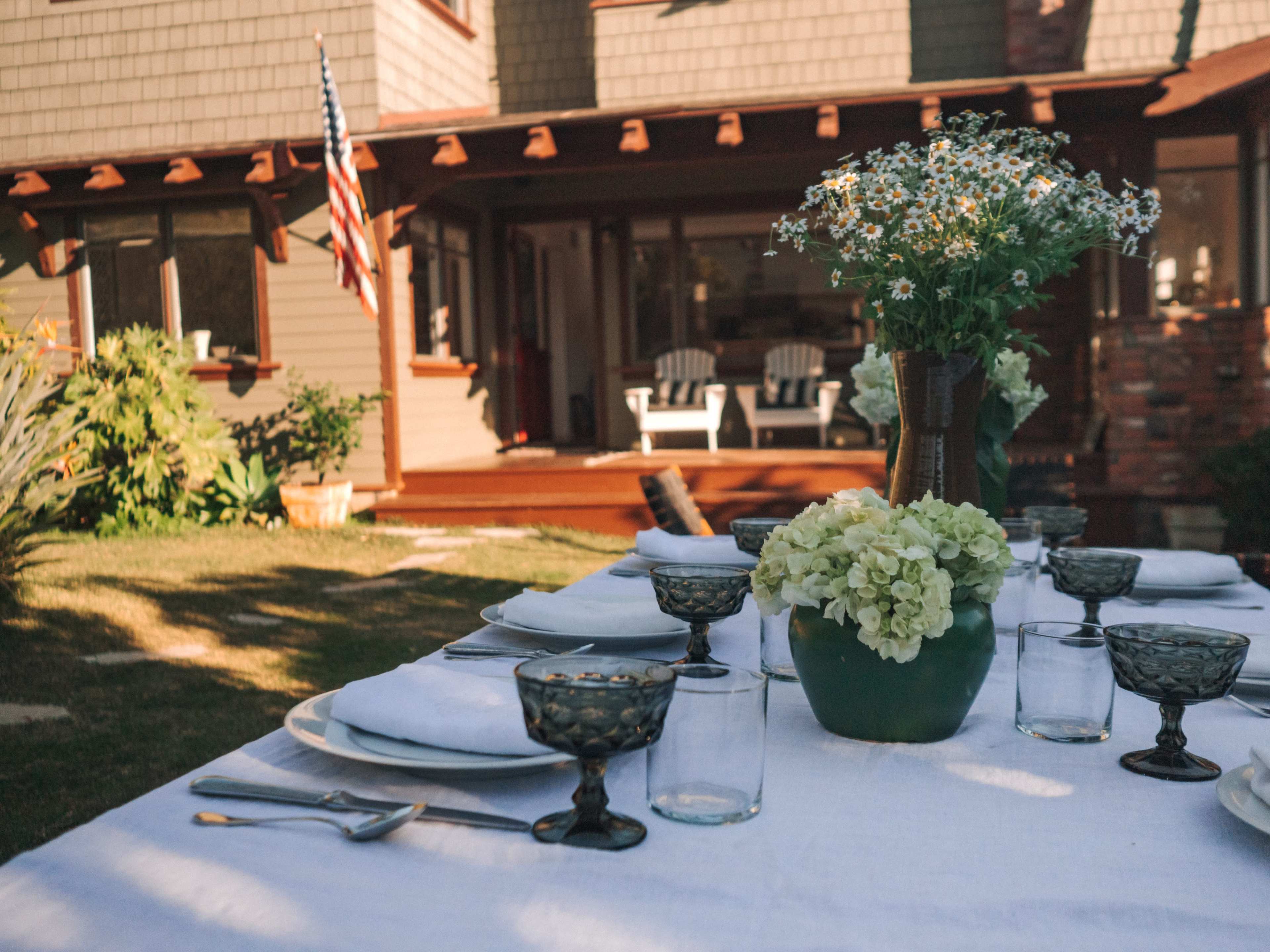 A beautifully set outdoor dining table is arranged with glassware and floral centerpieces in front of a house with a porch.