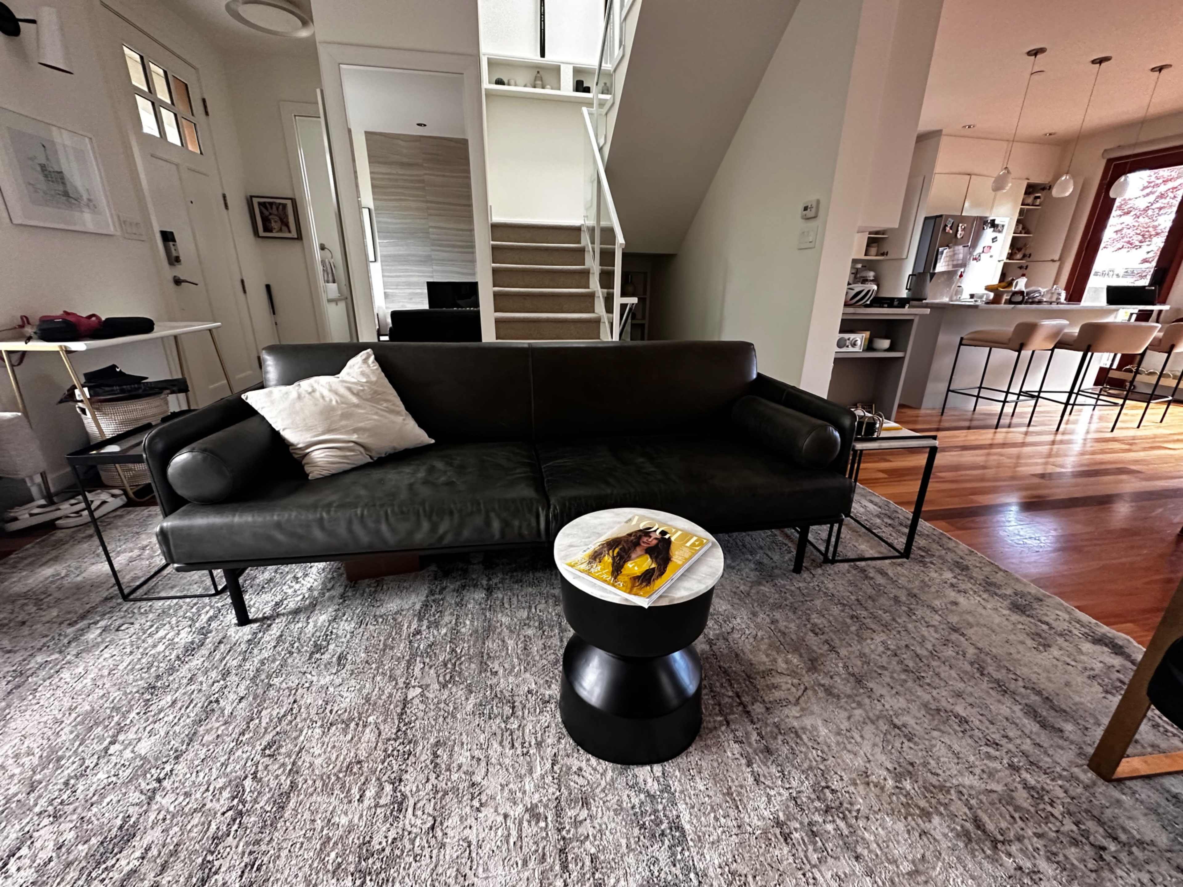 The image shows a modern living room with a black leather sofa, a round black side table with a yellow plate, and a staircase leading to an upper level.