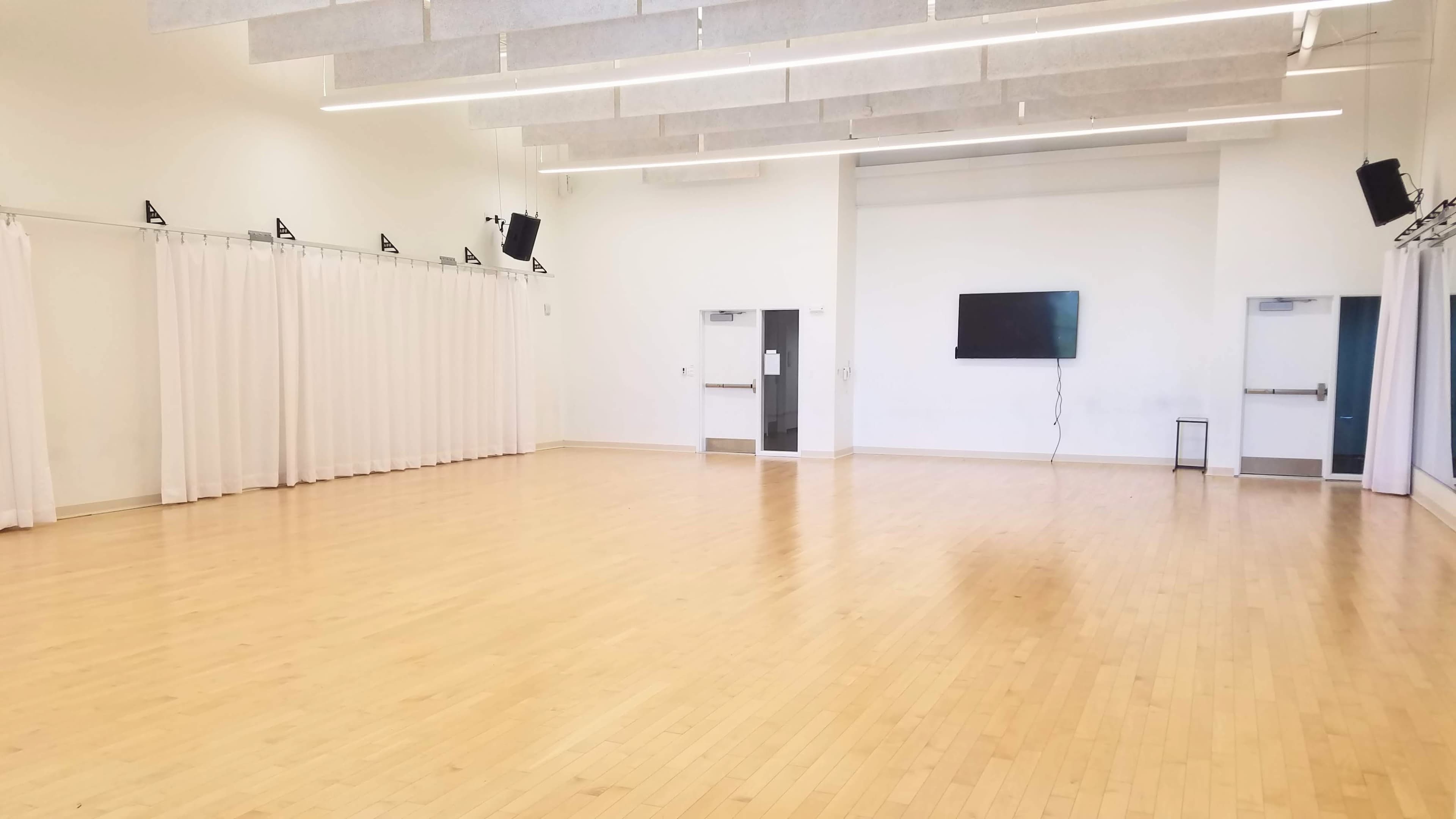 The image shows a spacious, empty studio with wooden flooring, white walls, and a television mounted on one wall.