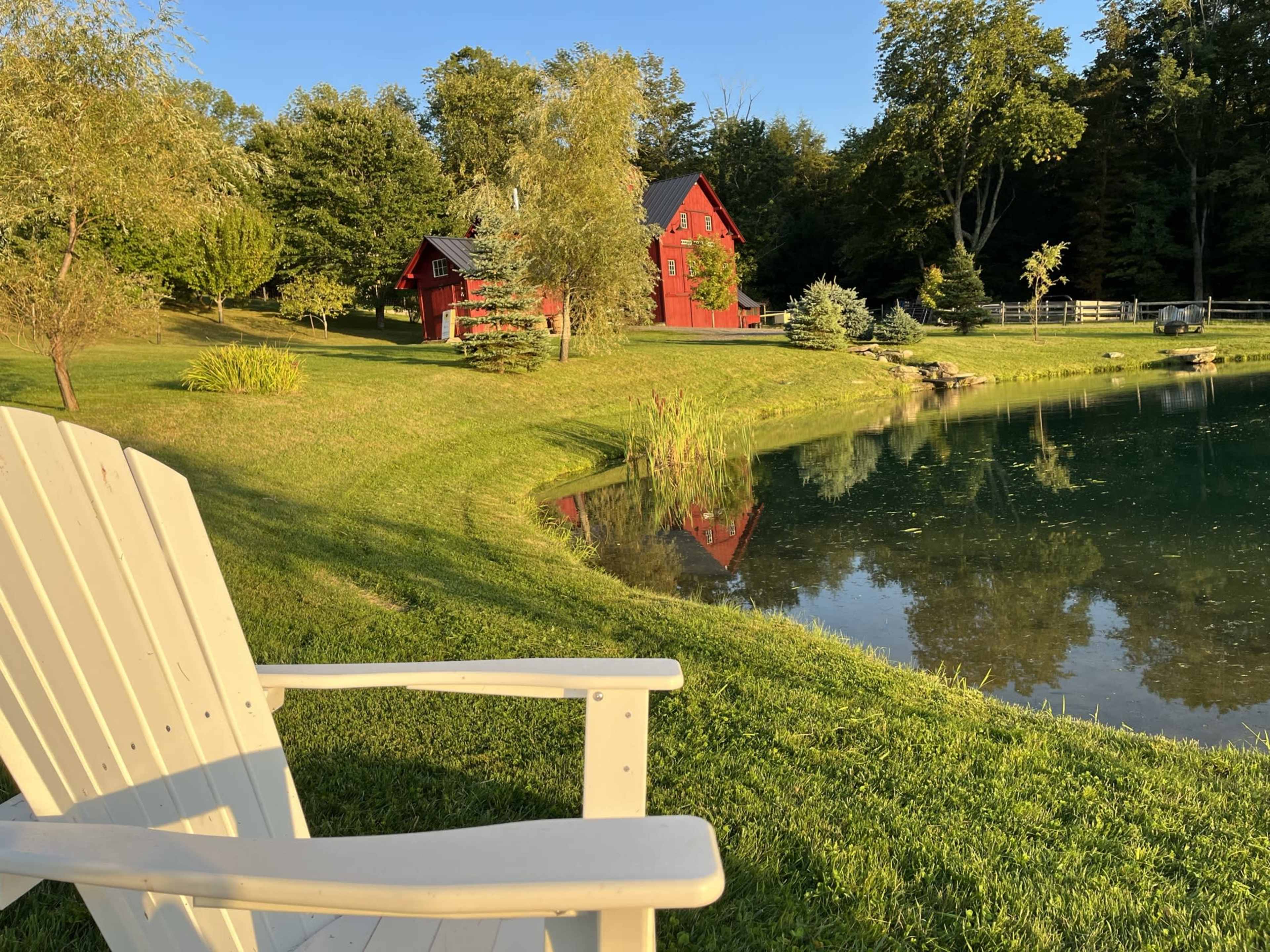 A white Adirondack chair sits in front of a serene pond, with a red house and lush greenery reflected in the water.