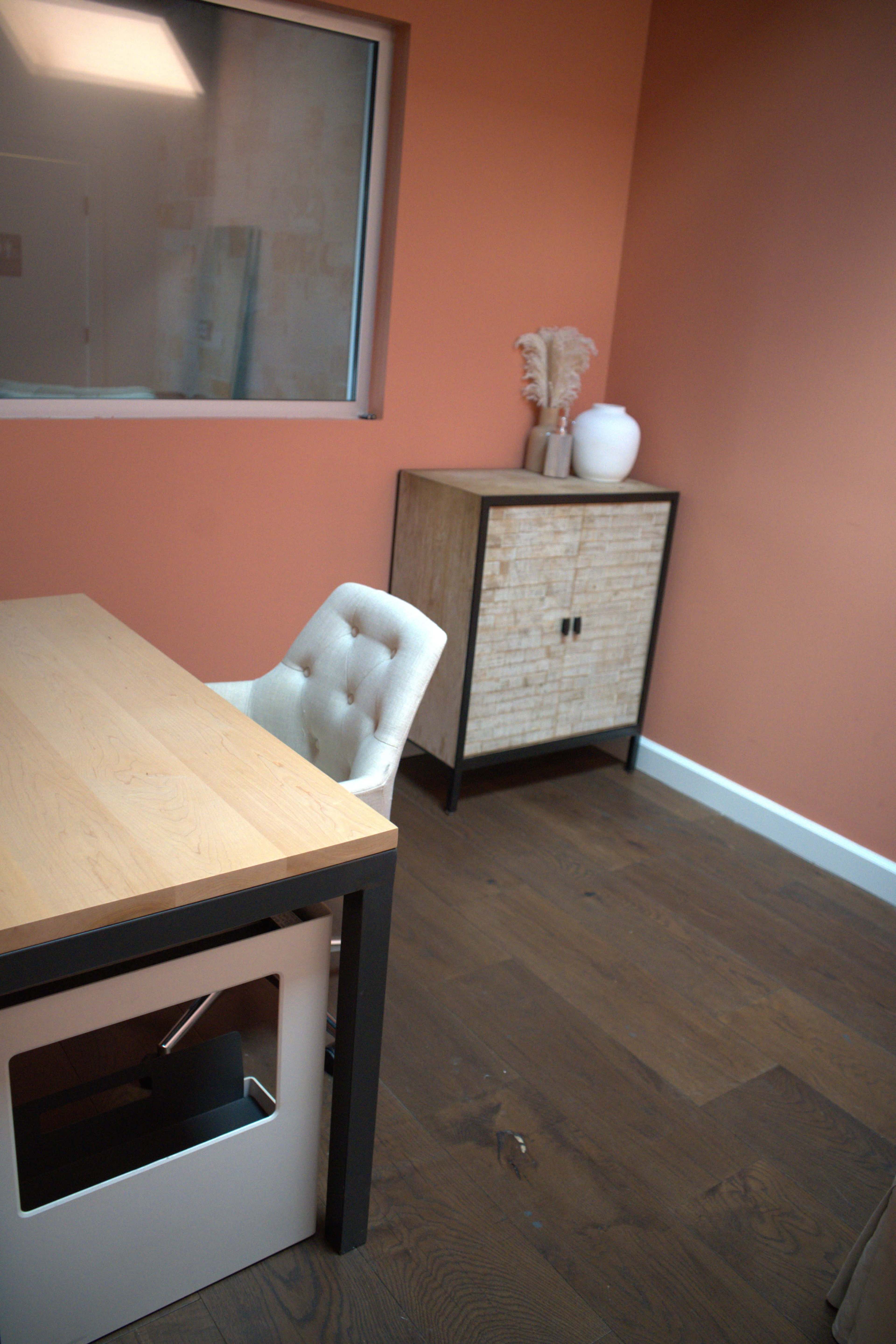 The image shows a modern office space with a wooden desk, an upholstered chair, and a cabinet against an orange wall.