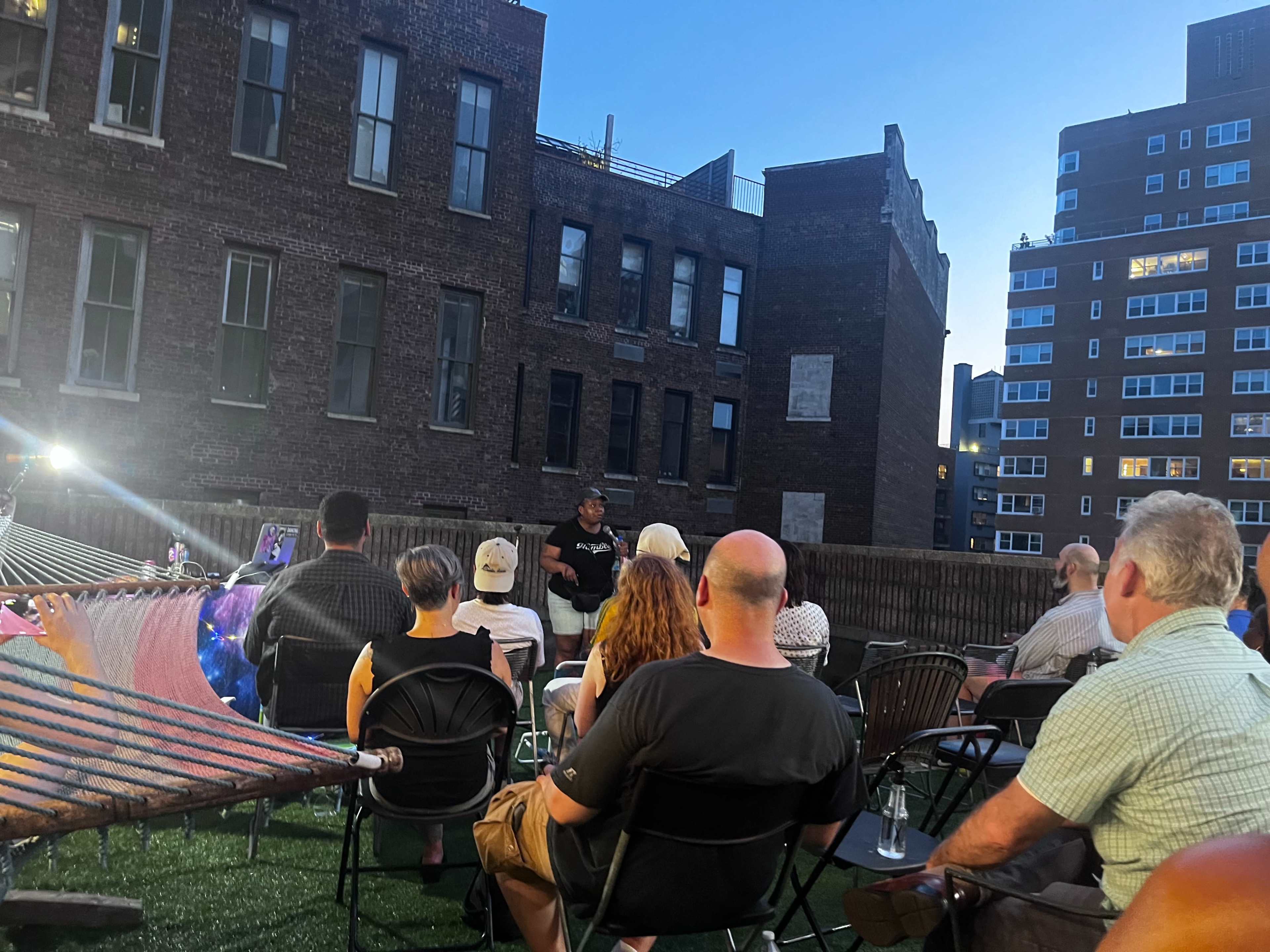 A group of people sits in folding chairs on a rooftop, facing a speaker in front of a brick building at dusk.