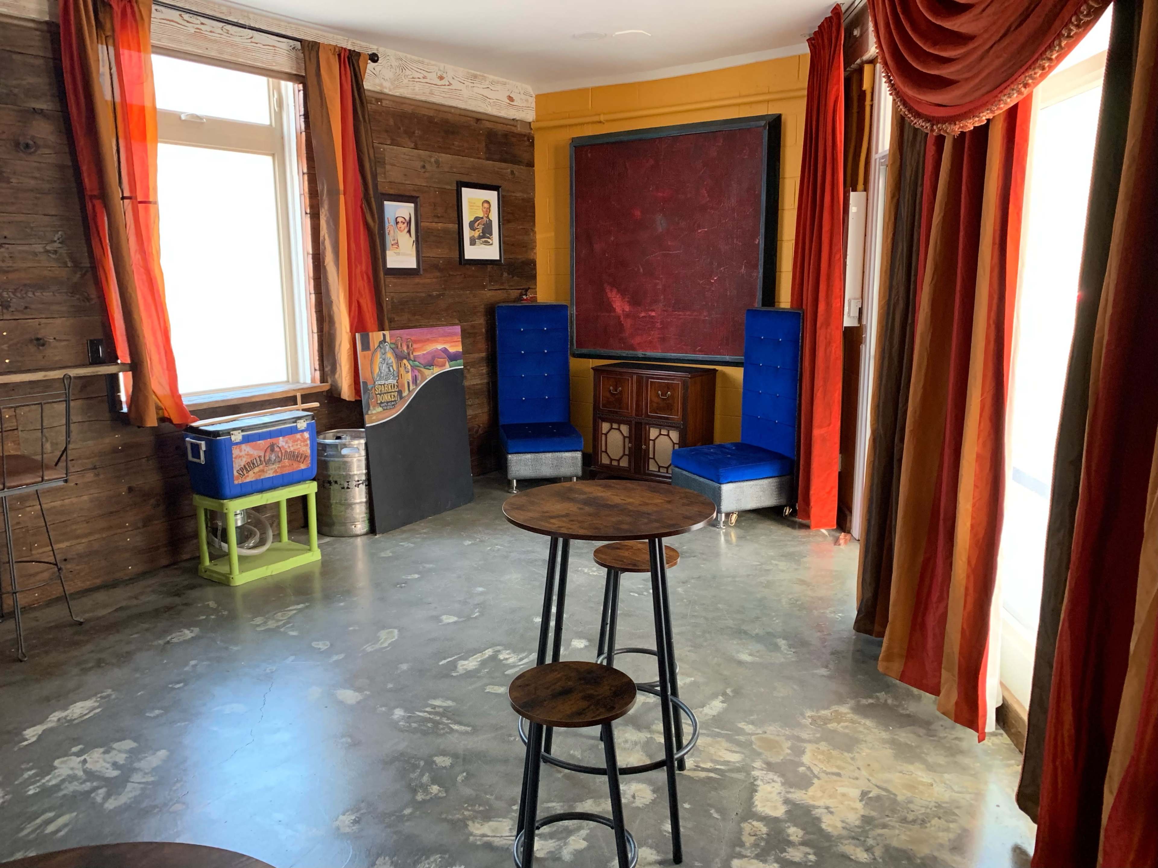 A room with a polished concrete floor, wooden walls, two blue upholstered chairs, and a small table set with three tall stools.