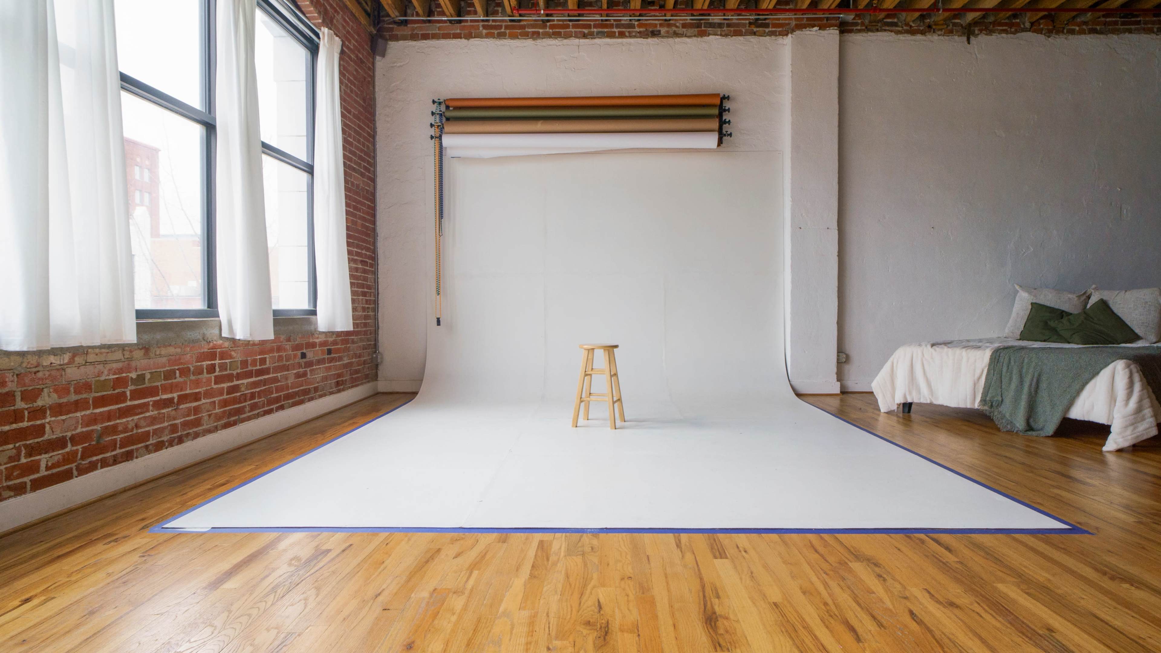 A wooden stool stands on a white backdrop in a bright, spacious room with large windows and exposed brick walls.