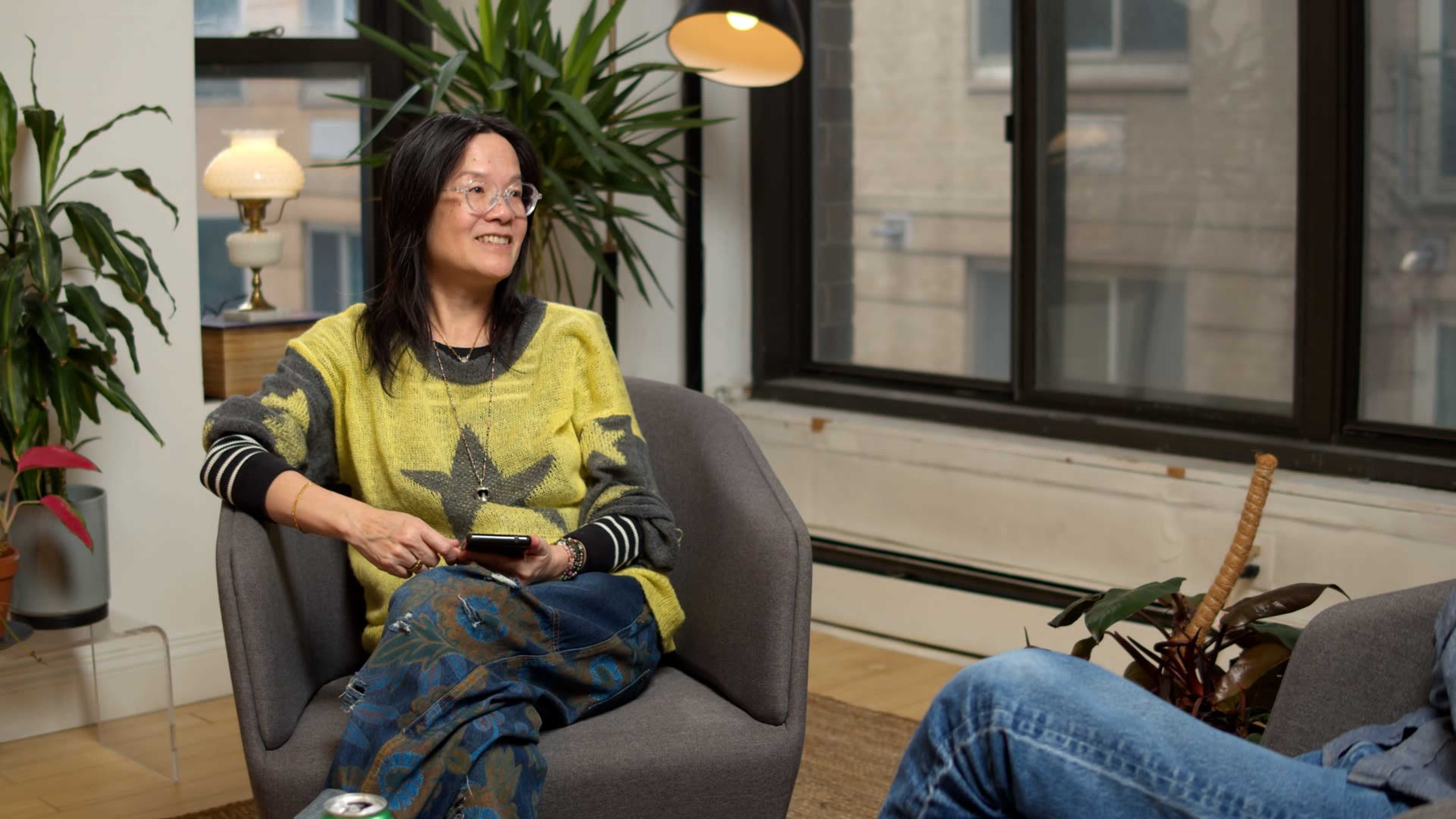 A person sits smiling in a cozy chair, wearing a colorful sweater and patterned pants, while engaged in conversation in a well-lit room with indoor plants.