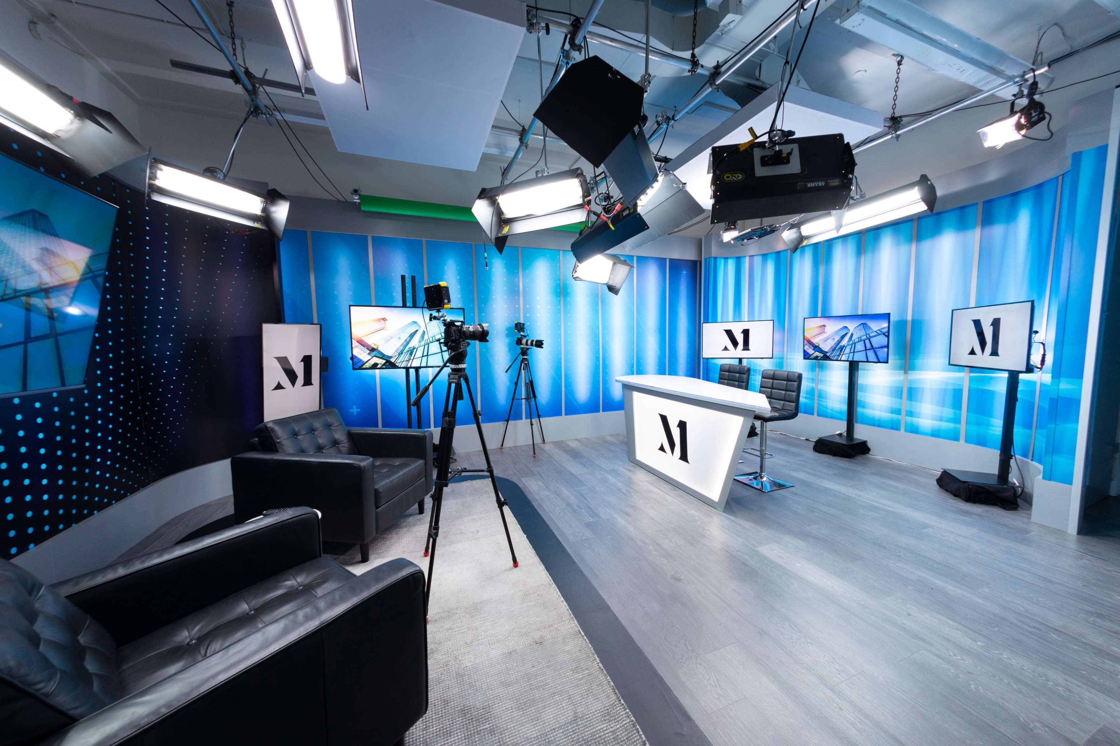 A modern studio setup with multiple cameras, lights, and a central desk surrounded by black leather chairs.