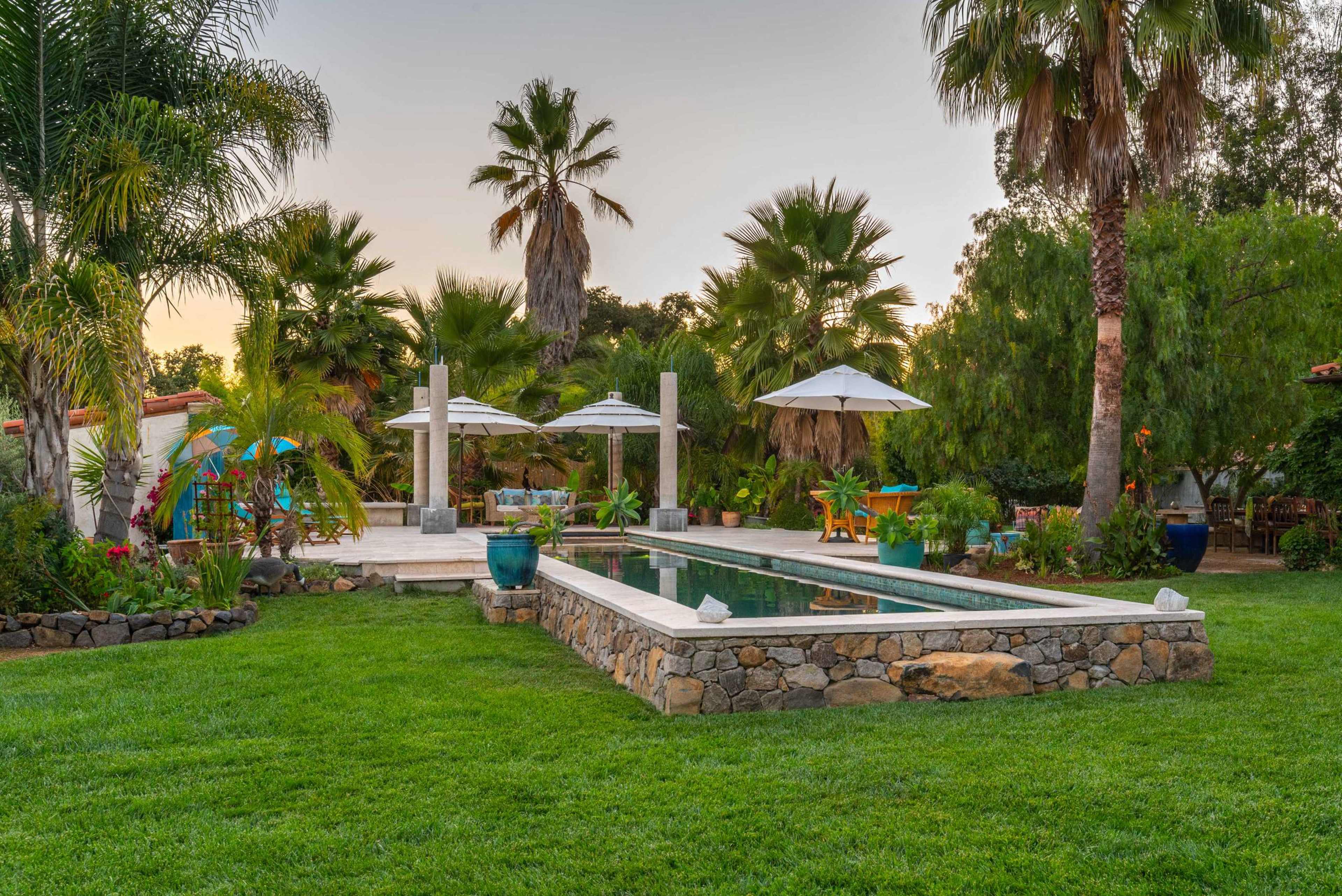 A landscaped backyard featuring a swimming pool, palm trees, and several shaded seating areas.