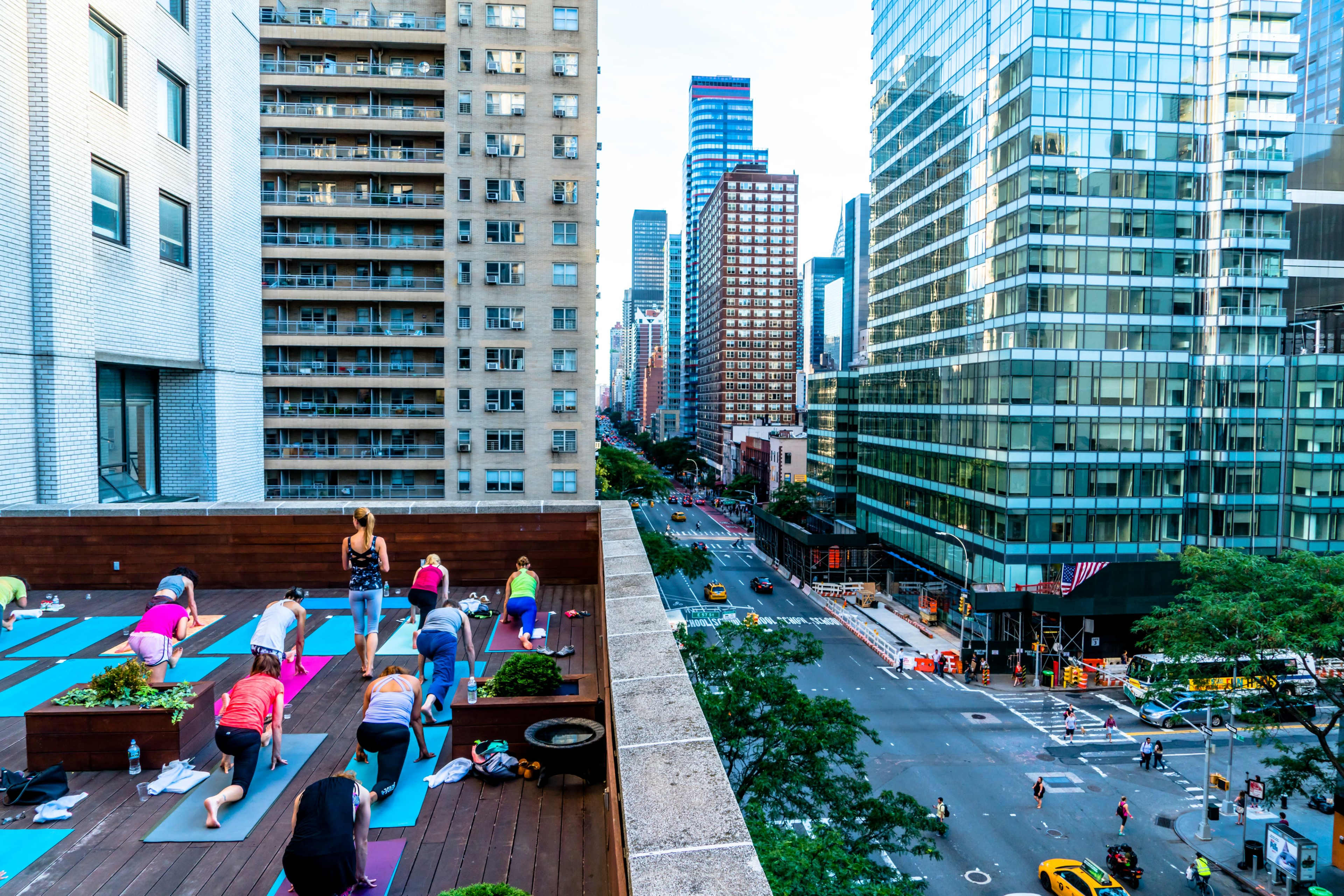 A group of people practices yoga on a rooftop in a city, with tall buildings and a street visible in the background.