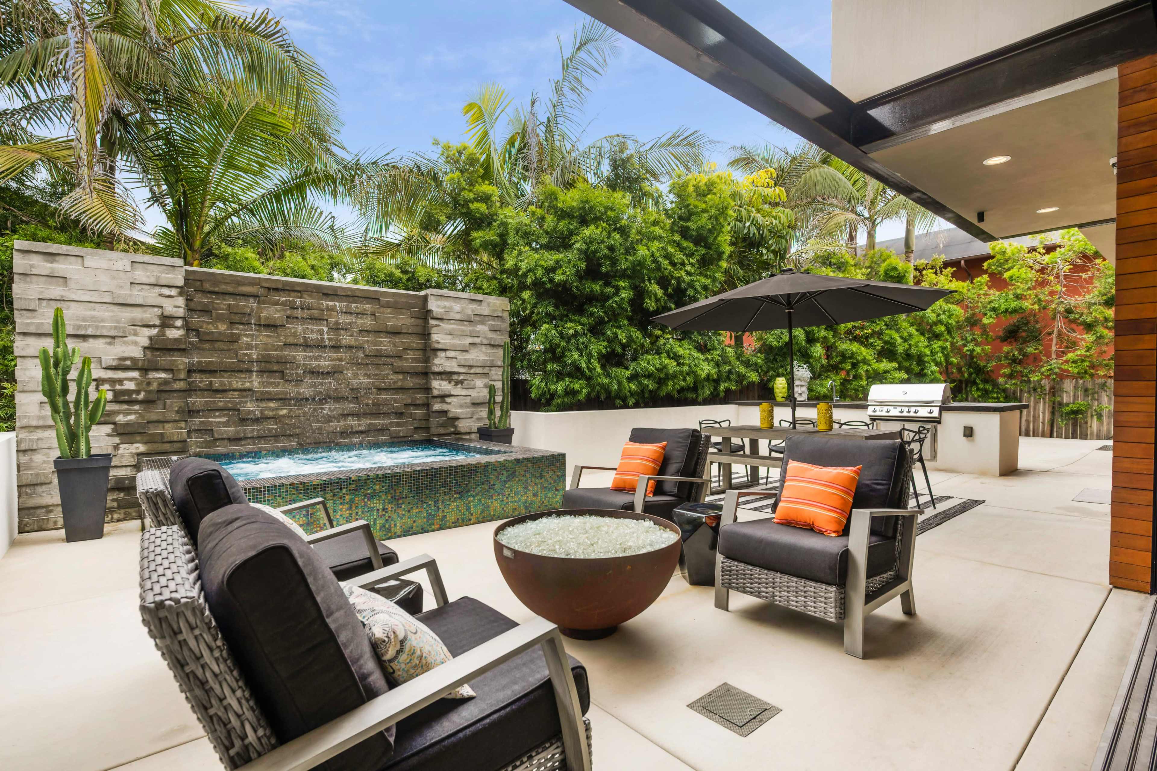 A modern outdoor patio area featuring a water fountain, seating with cushions, a dining table under an umbrella, and a built-in grill surrounded by lush greenery.