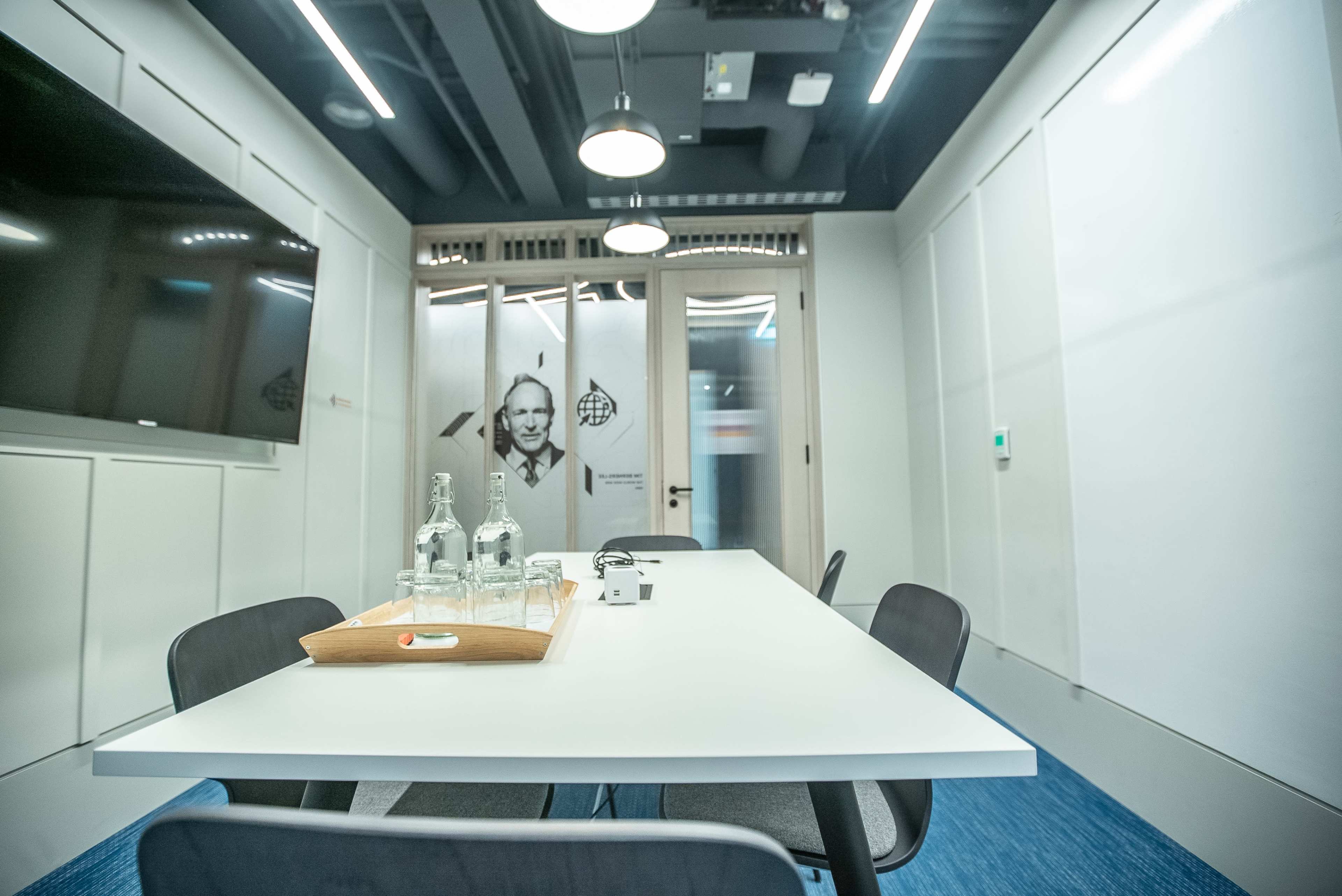 A modern conference room features a long white table, chairs, water bottles, and a large television screen on the wall.
