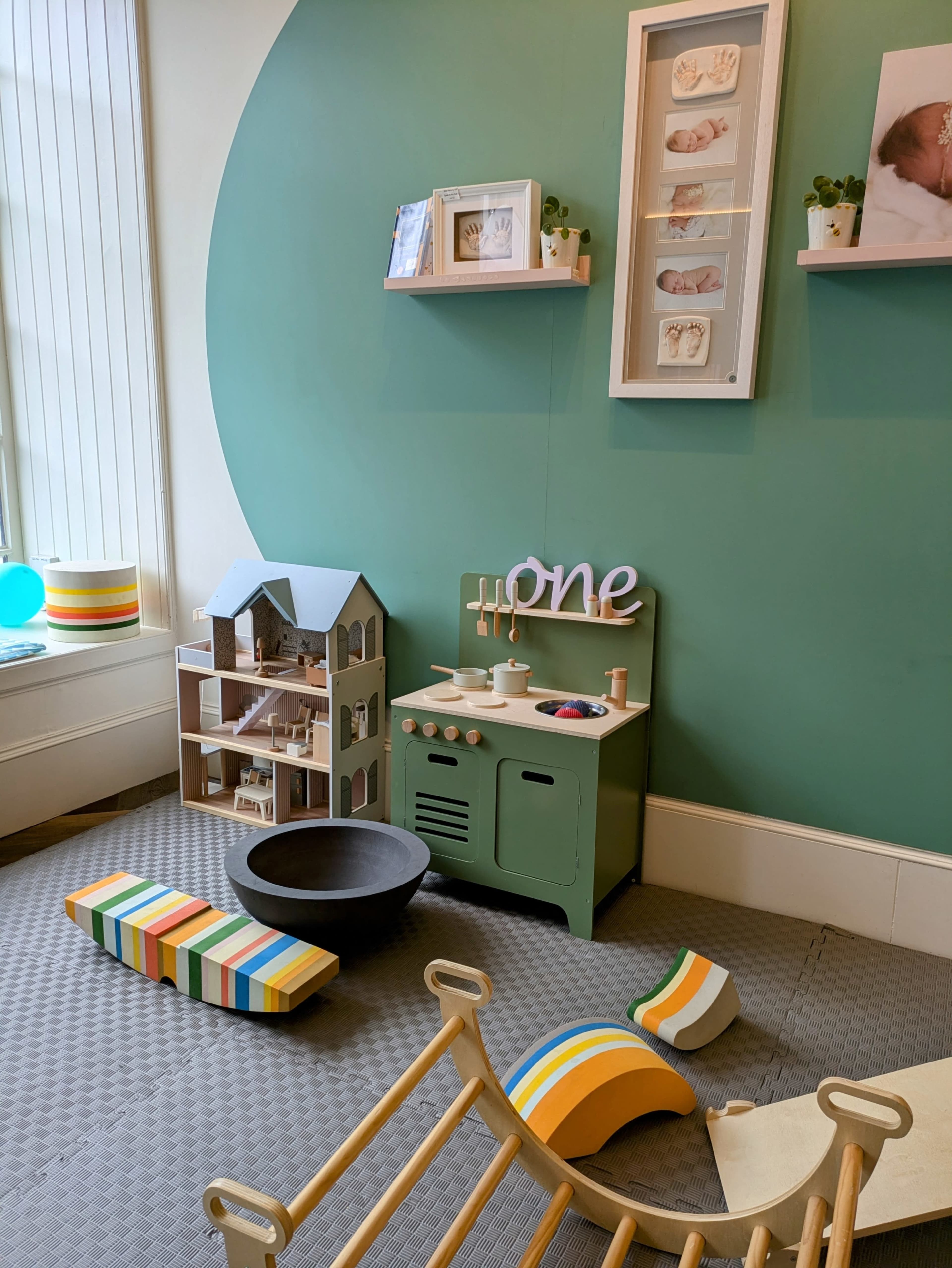 A playful corner with a green wall featuring a small kitchen set, a dollhouse, and various colorful toys scattered on the floor.