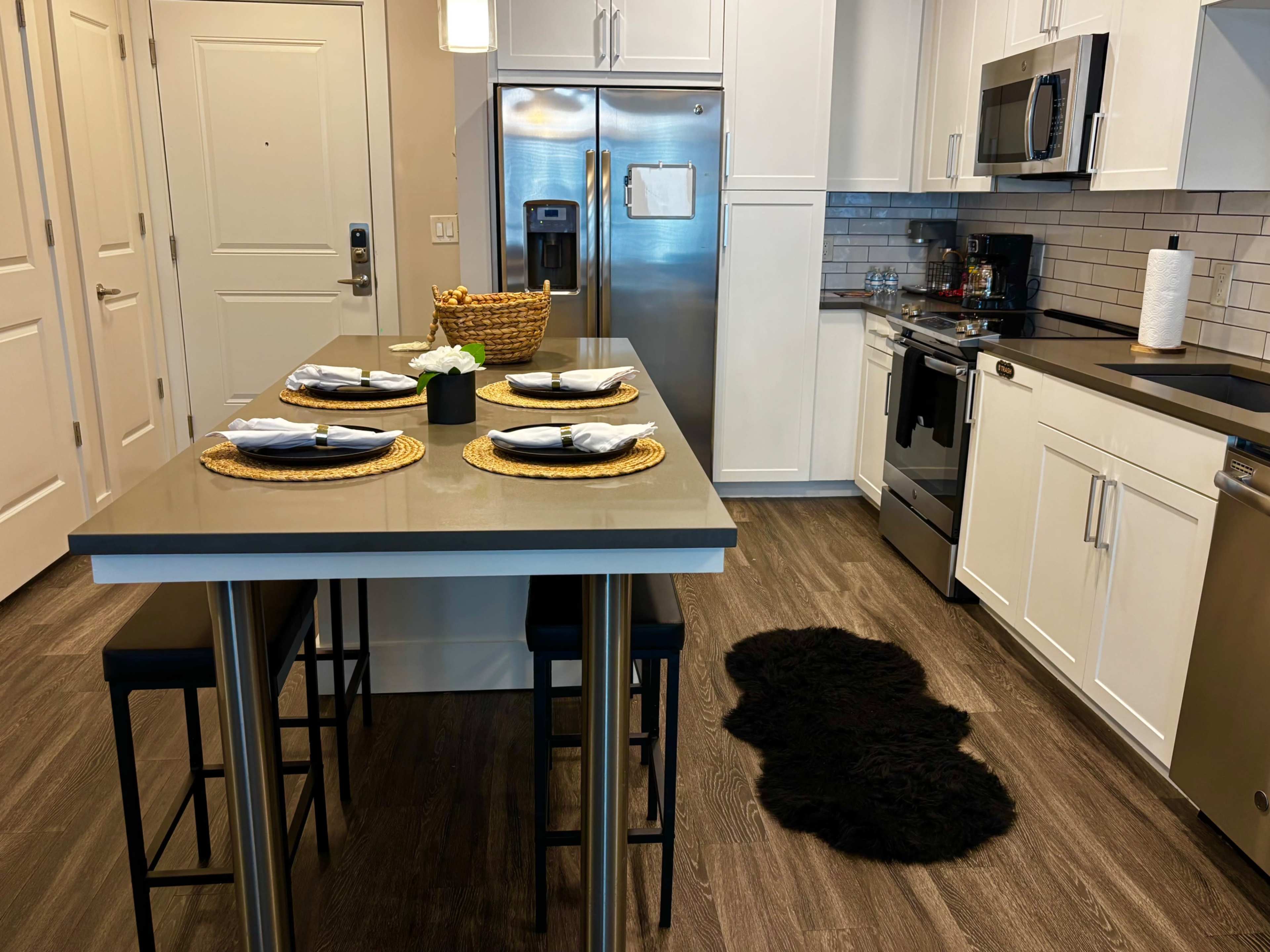 The image shows a modern kitchen with a dining island featuring four place settings, stainless steel appliances, and light-colored cabinetry.