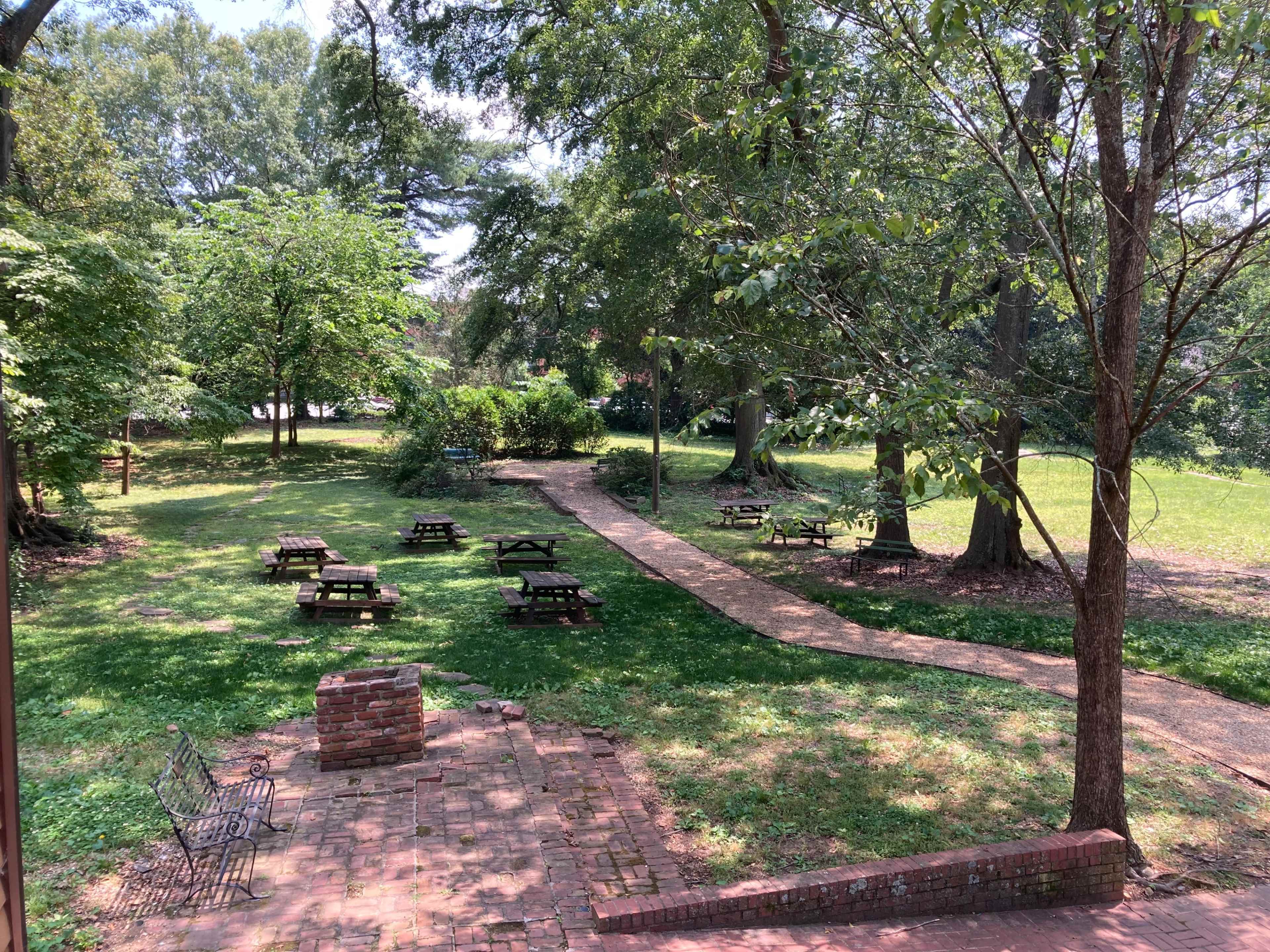 The image shows a park area with several picnic tables arranged on a grassy field, bordered by trees and a winding path.