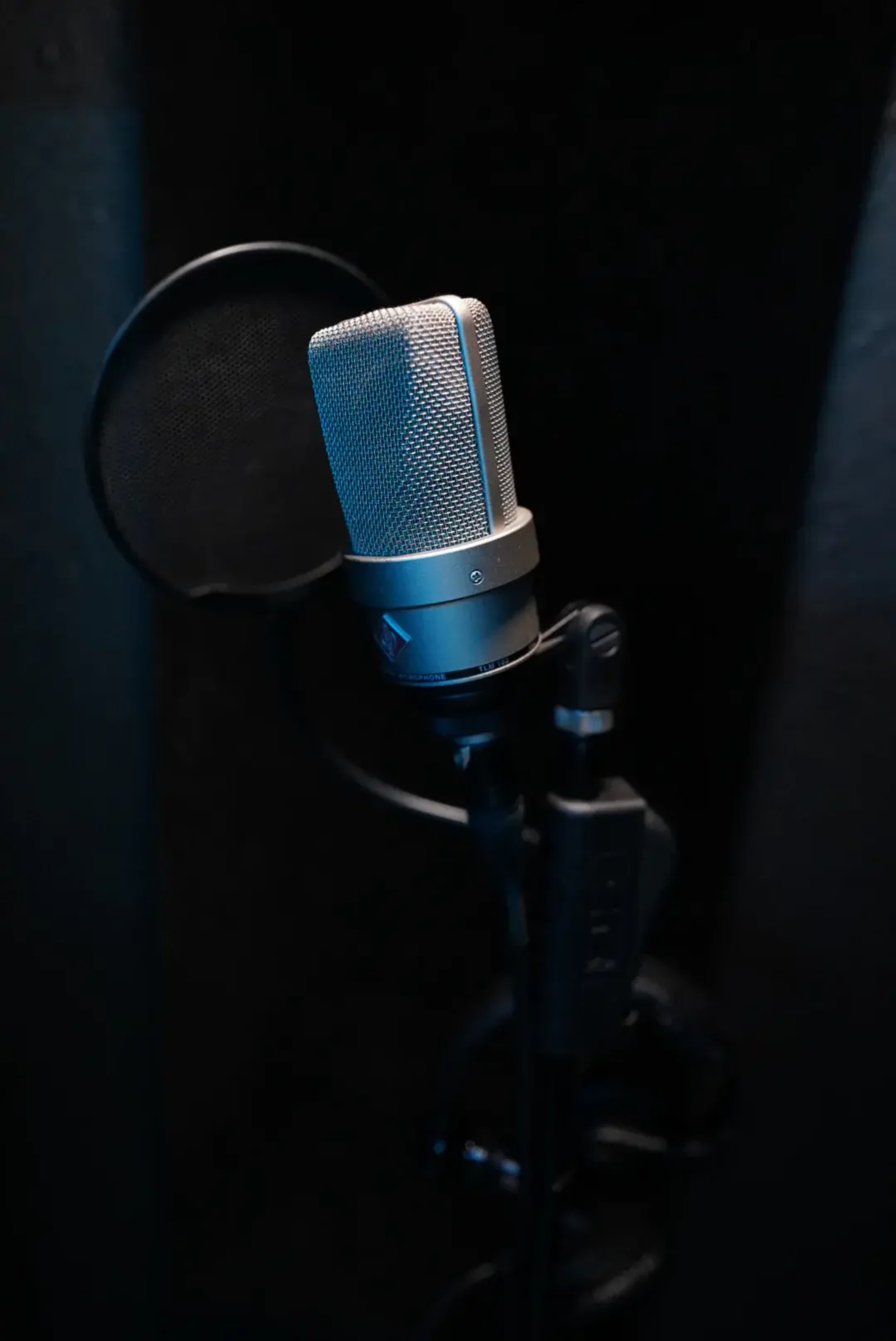 A silver microphone stands mounted on a black microphone stand in a dimly lit sound booth.