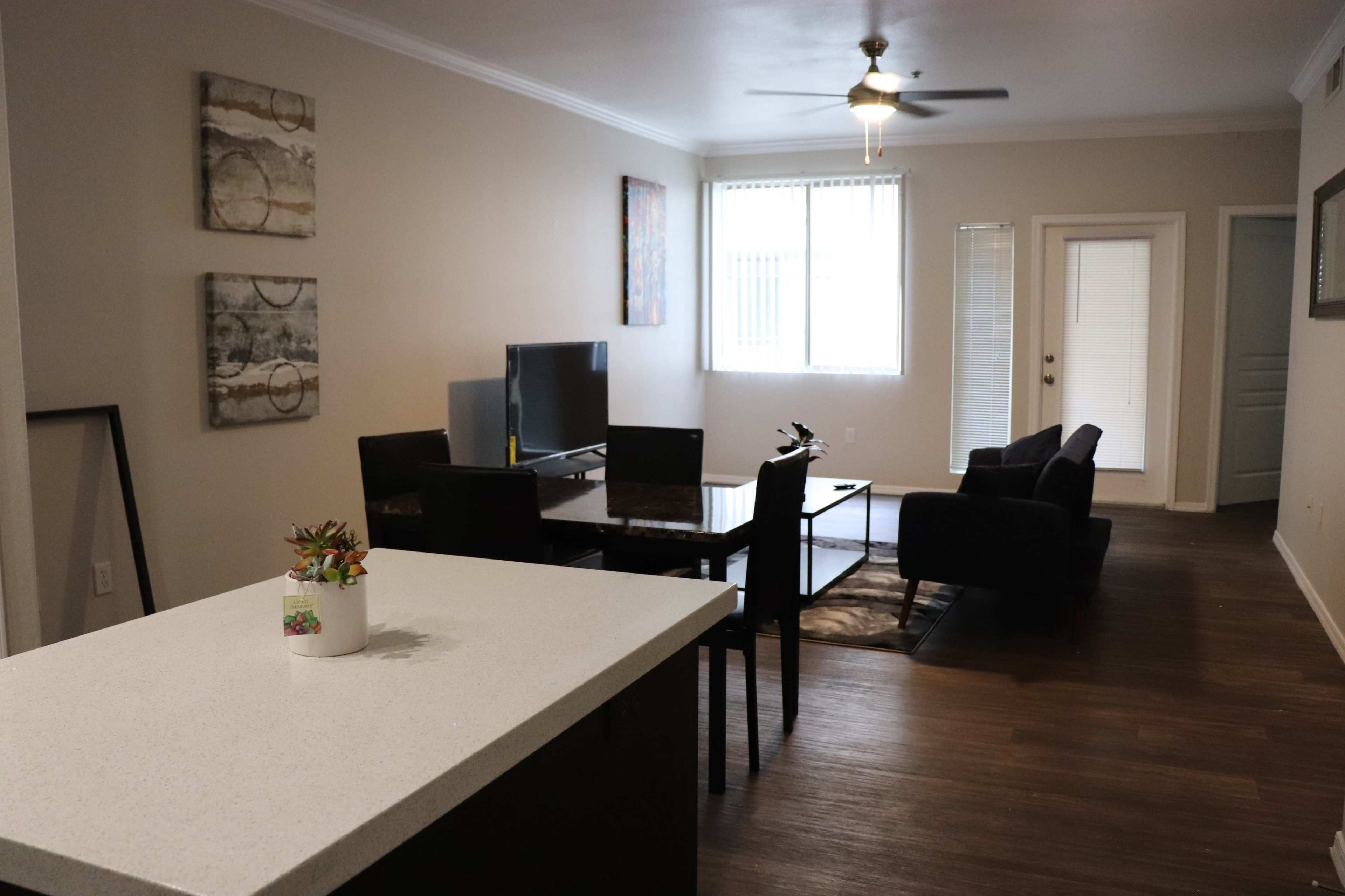 The image shows a modern living room and dining area with a television, dining table, and seating arrangement, viewed from a kitchen counter.