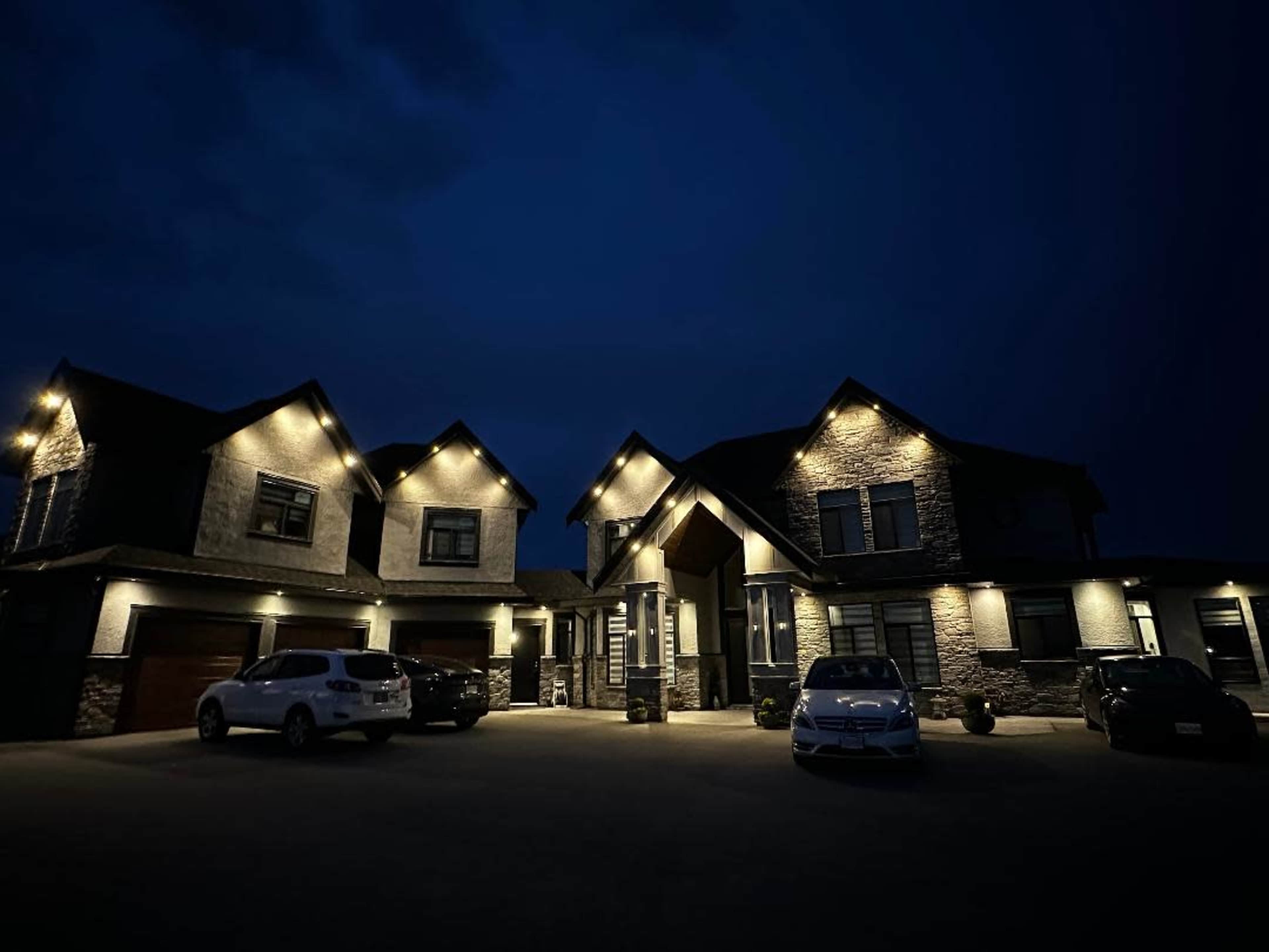 A modern two-story house with stone accents and decorative lighting is situated at night, surrounded by several parked cars in a well-lit driveway.