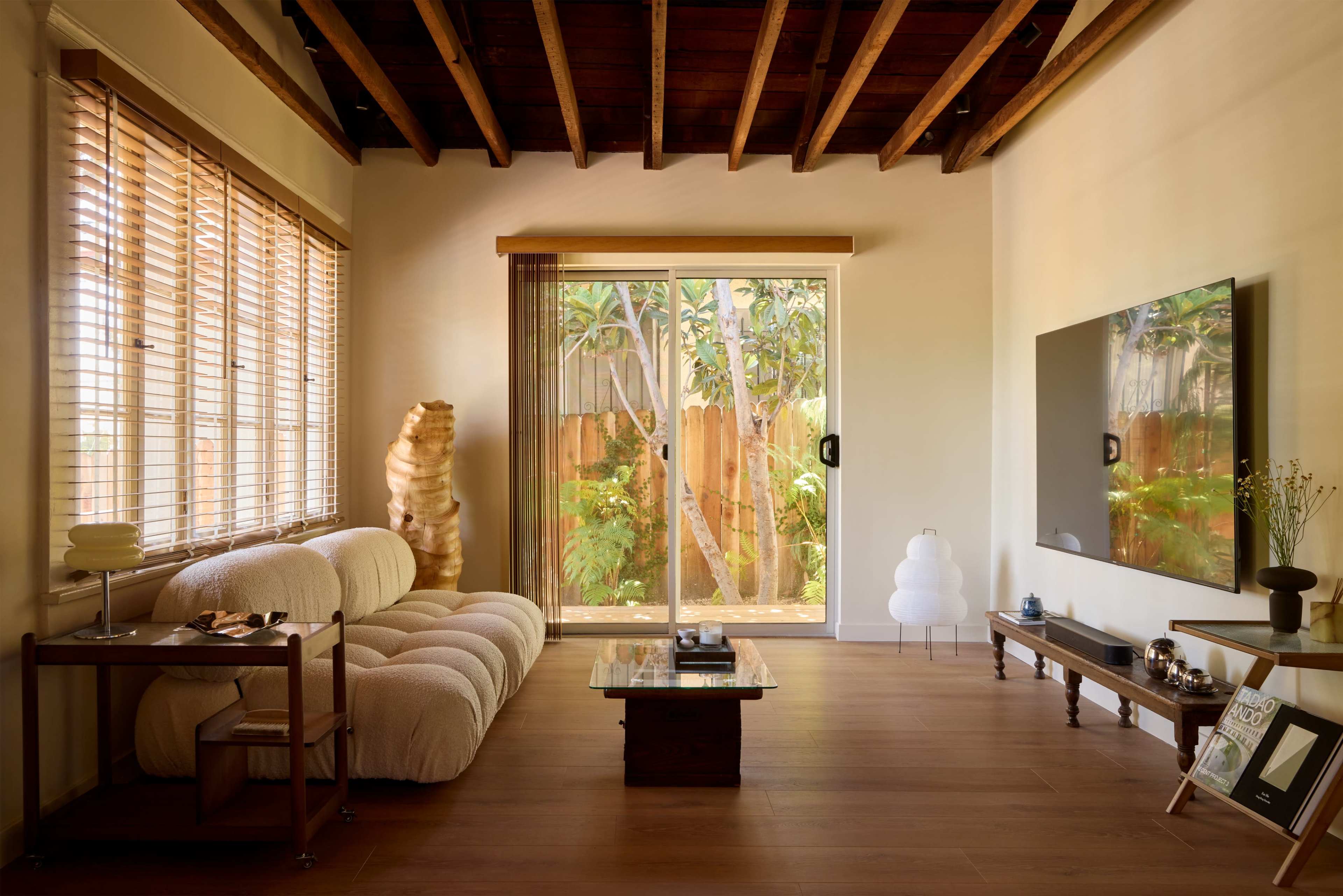 The image depicts a modern living room with a large window opening to a garden, featuring a plush sofa, a wooden coffee table, and a television mounted on the wall.