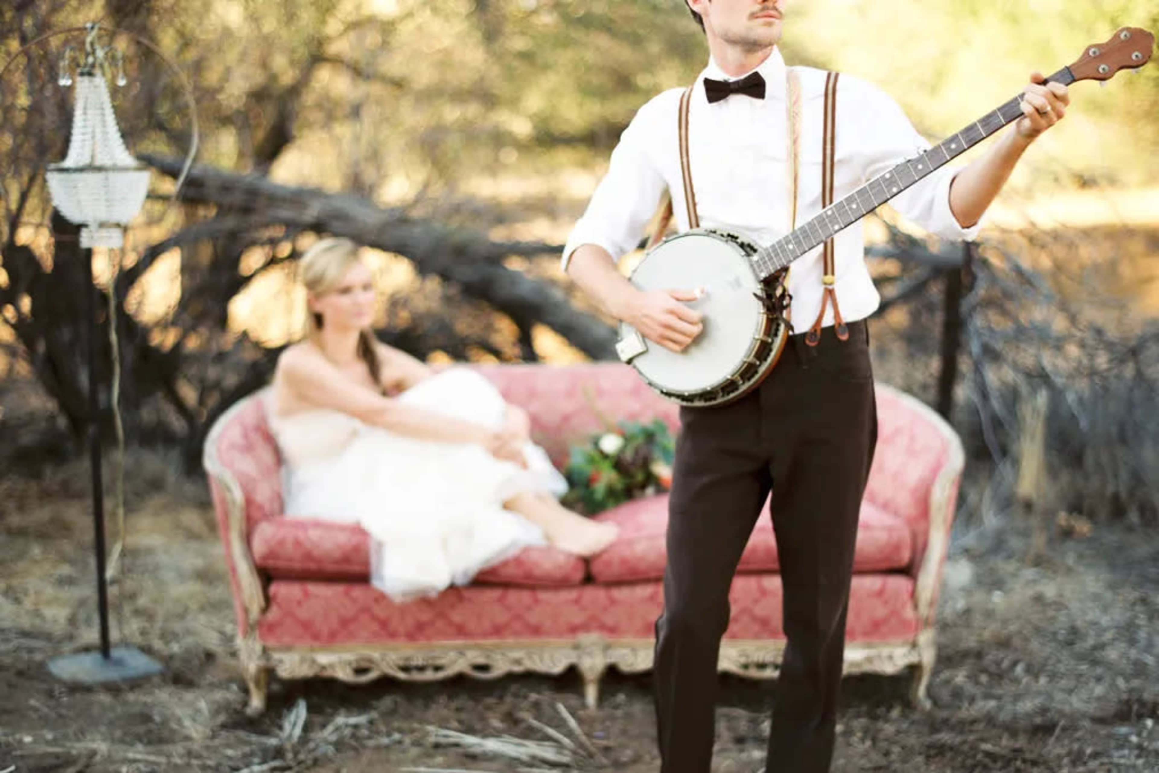A man in formal attire plays a banjo while a woman in a white dress sits on a vintage sofa in a rural setting.