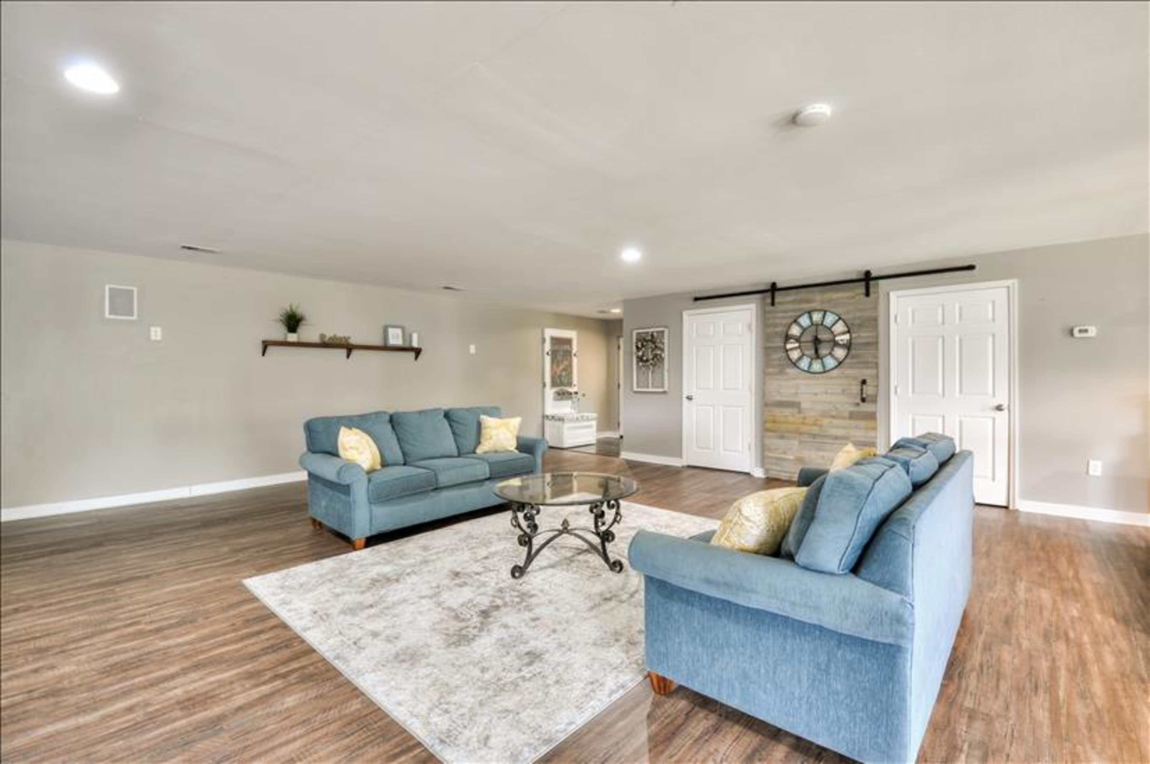 A spacious living room featuring two blue sofas, a glass coffee table, and a decorative wall clock against a wood-paneled accent wall.