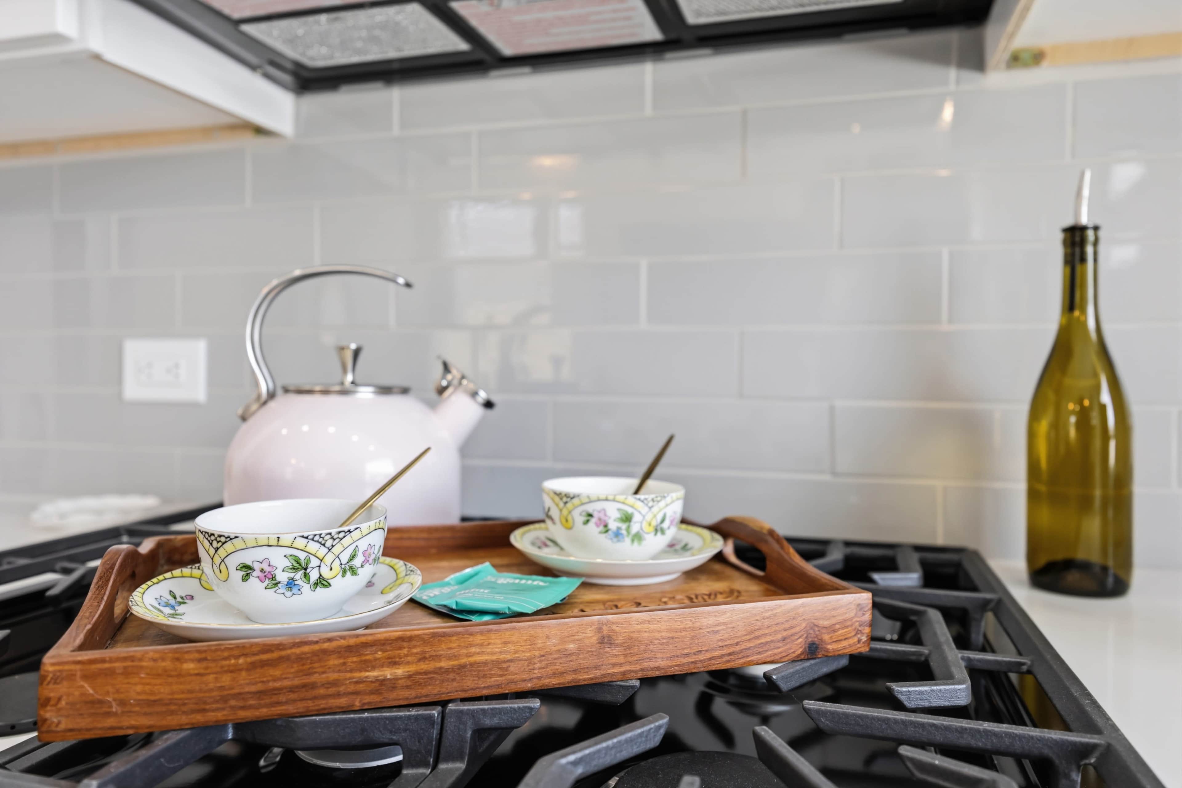 A wooden tray holds two decorated teacups and a small packet, next to a pink kettle and an olive oil bottle on a gas stove.