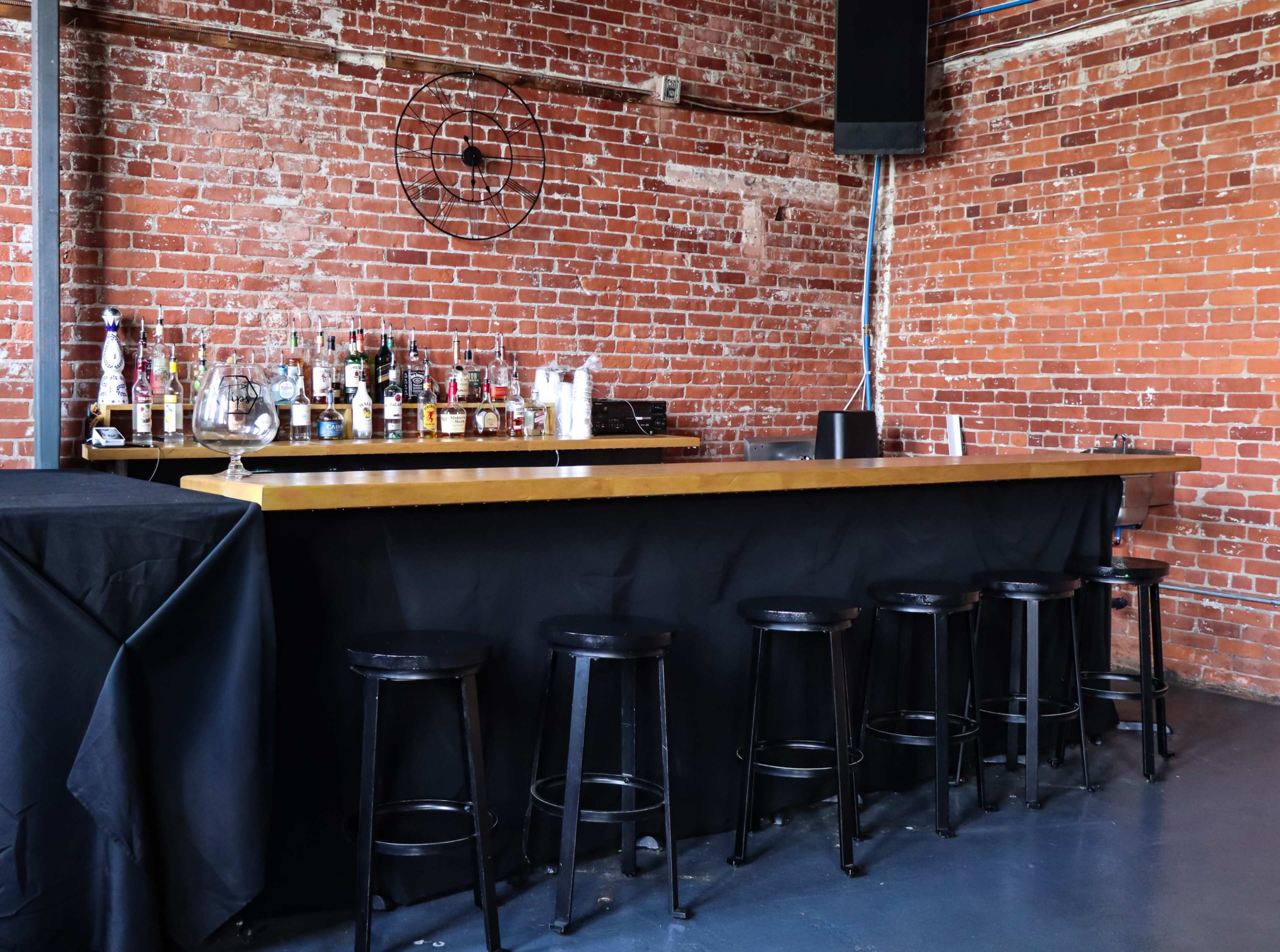A wooden bar with high black stools is positioned against a brick wall, with various bottles displayed on the bar and a clock mounted on the wall.