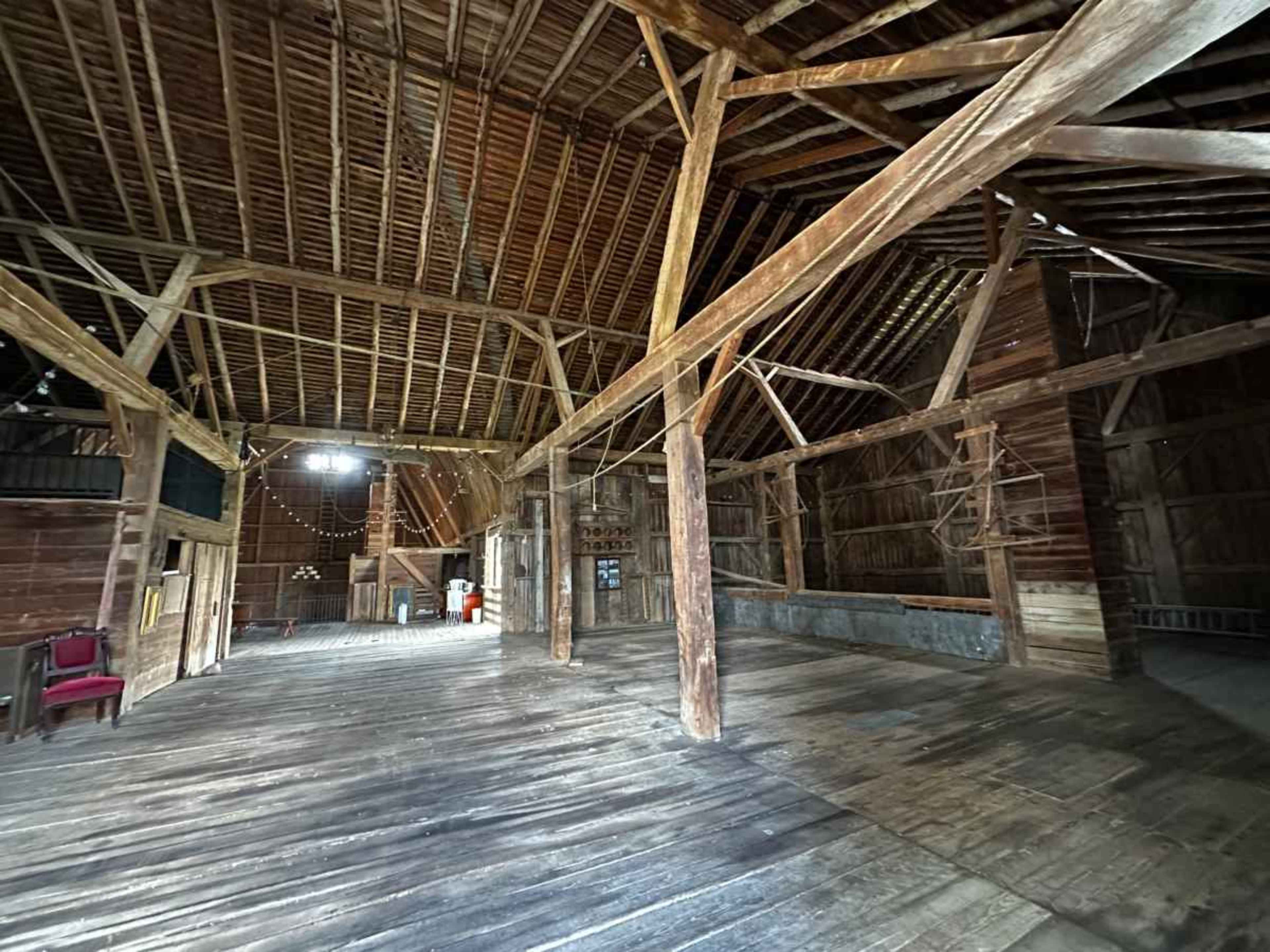The image shows a spacious, wooden barn interior featuring exposed beams, a wooden floor, and large open areas.