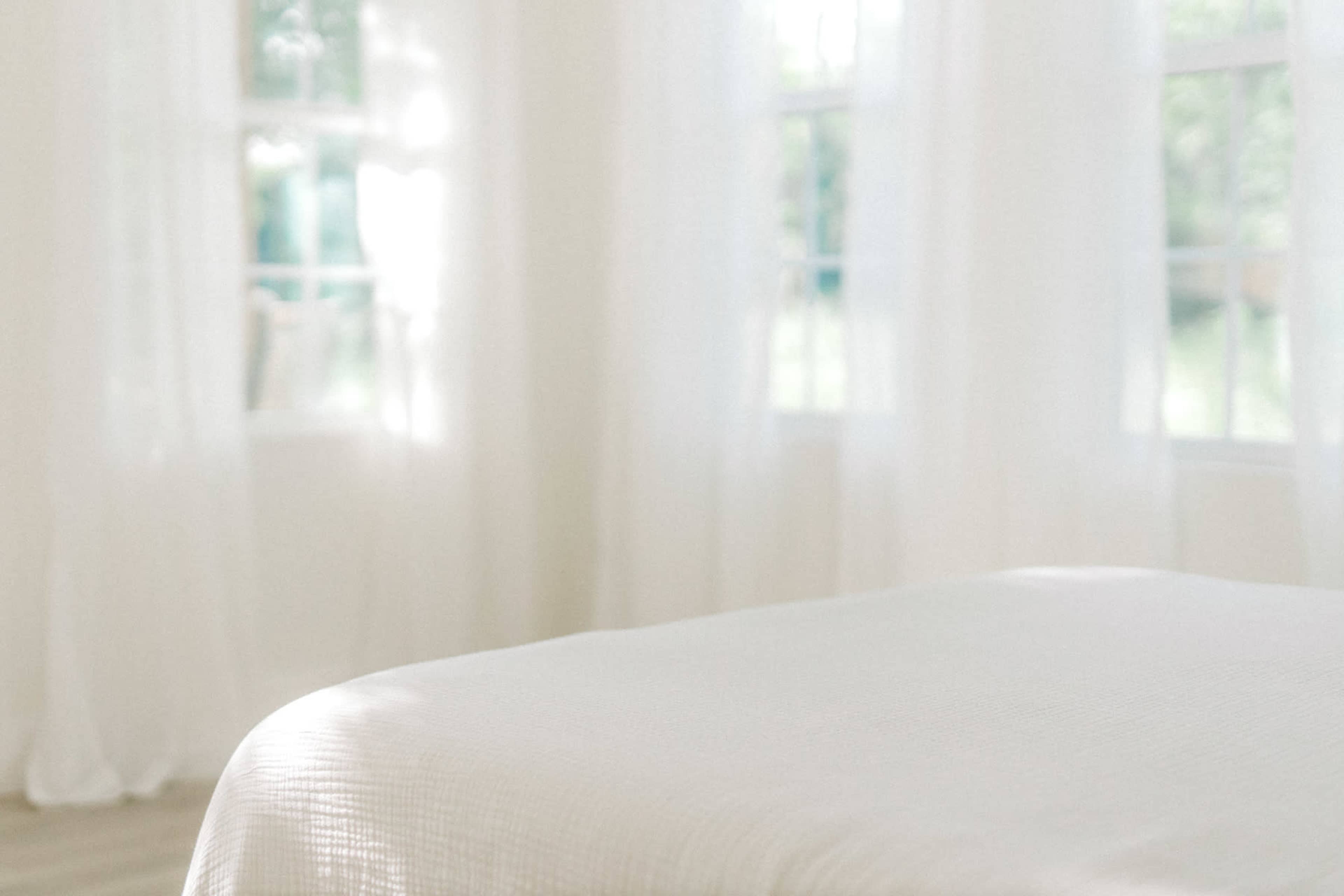 A softly lit room features a white bedspread and sheer curtains letting in natural light.