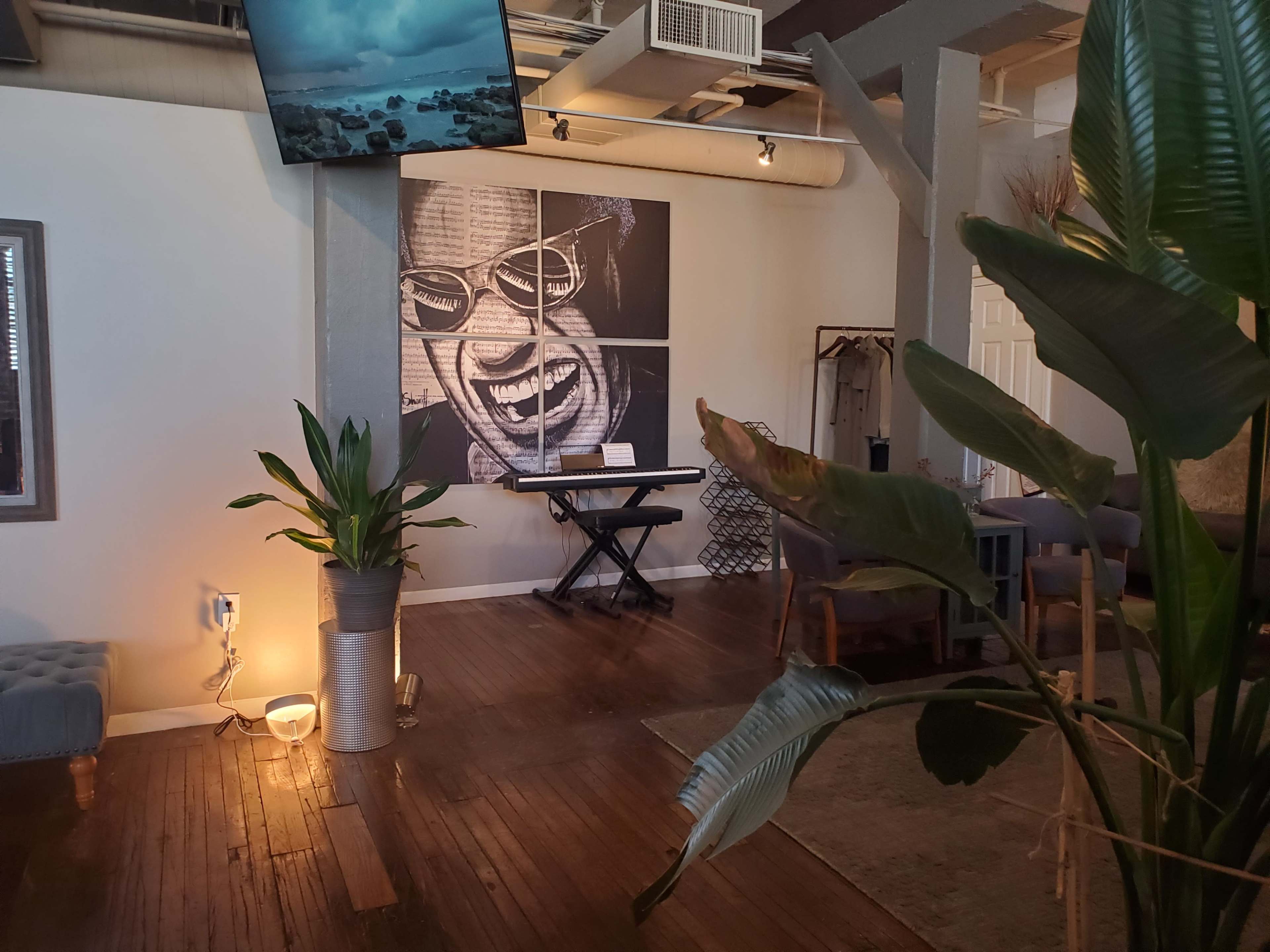 The image shows an interior space featuring a large mural of a smiling face, a piano, and plants, with seating arranged around the area.