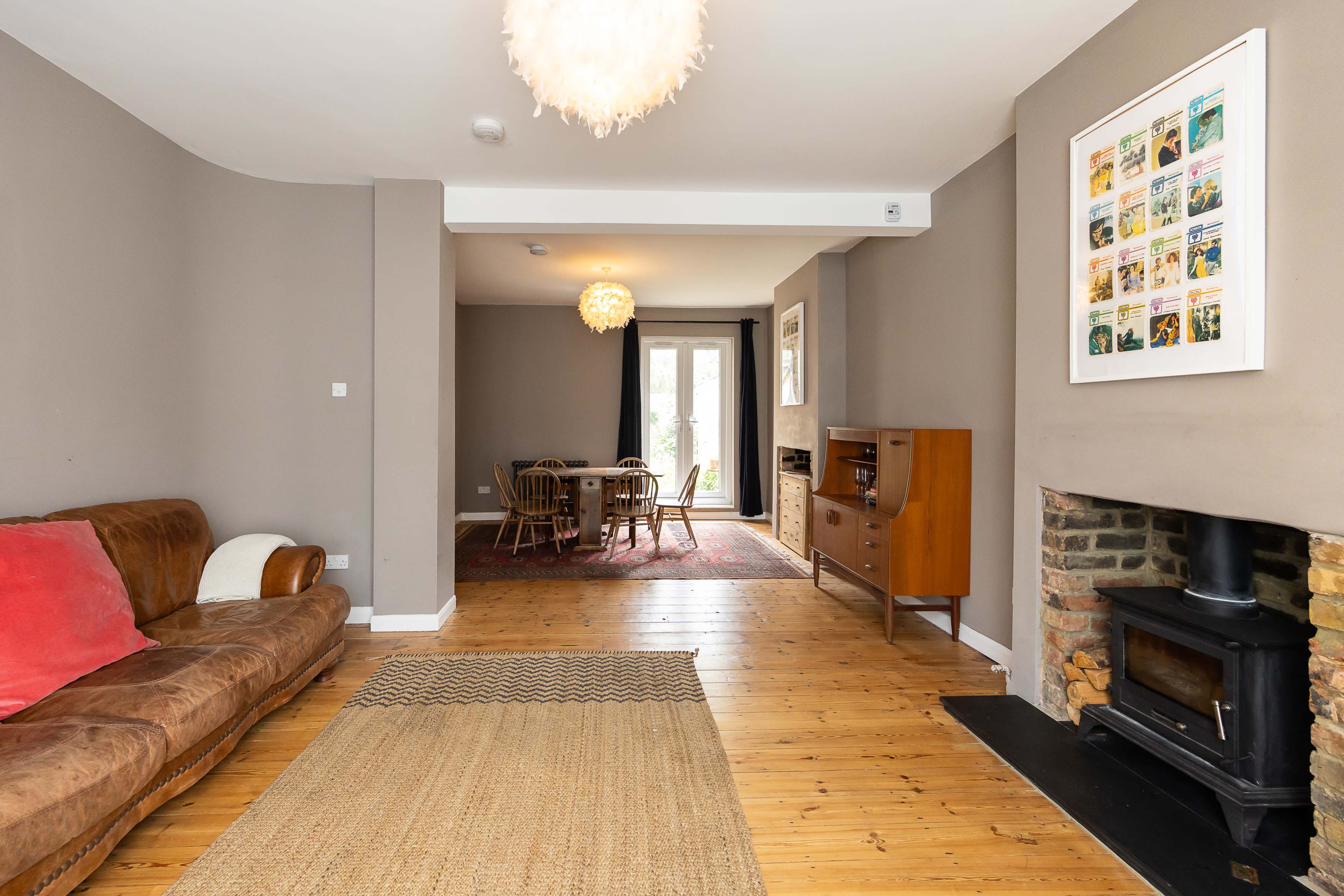 The image shows a living room with a brown leather sofa, a woven rug on wooden flooring, and a dining area in the background that features a table with chairs and a fireplace.