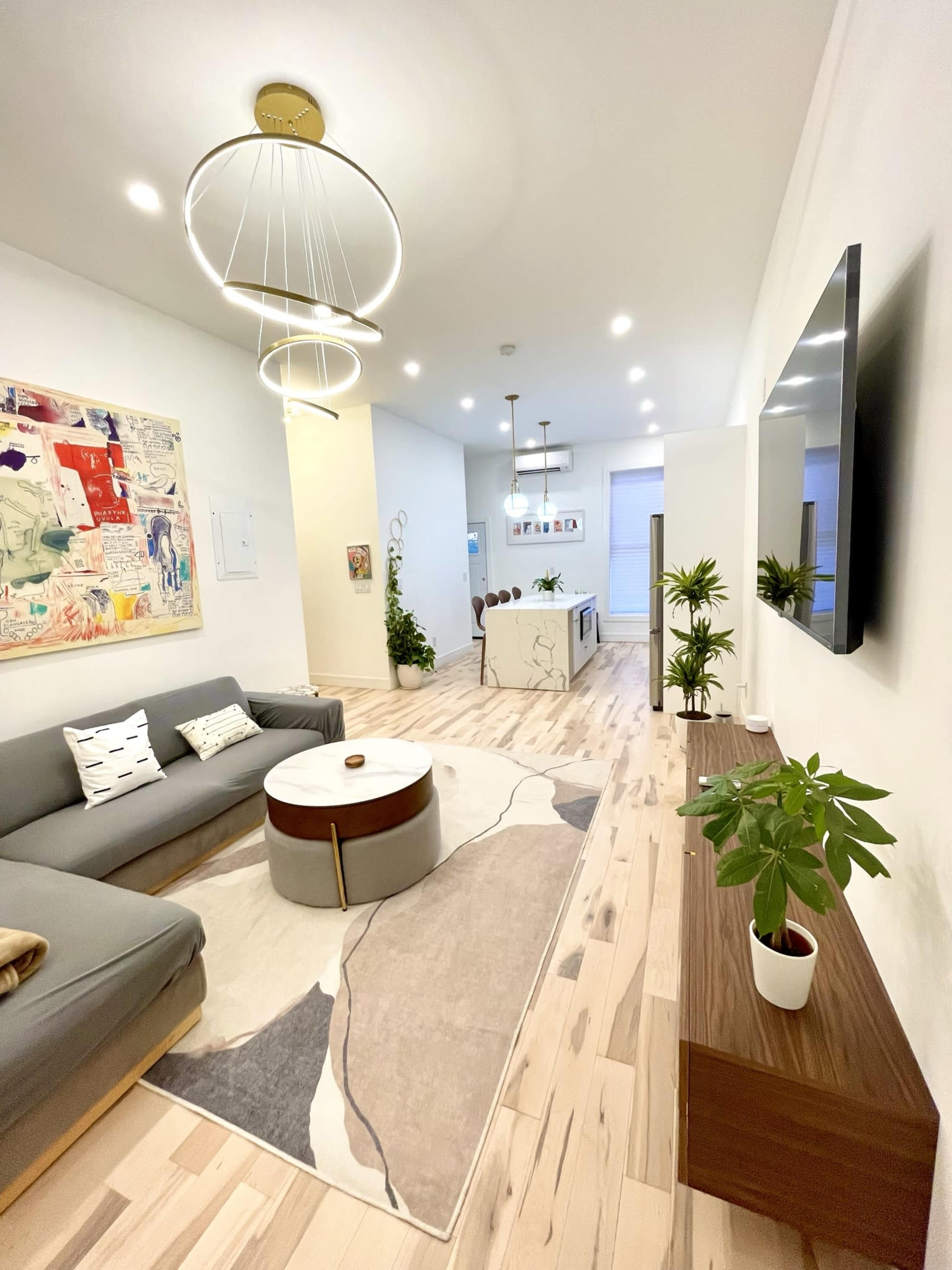 A modern living room features a gray sofa, a round coffee table, plants, and two circular light fixtures, with a large wall-mounted television and an open kitchen visible in the background.