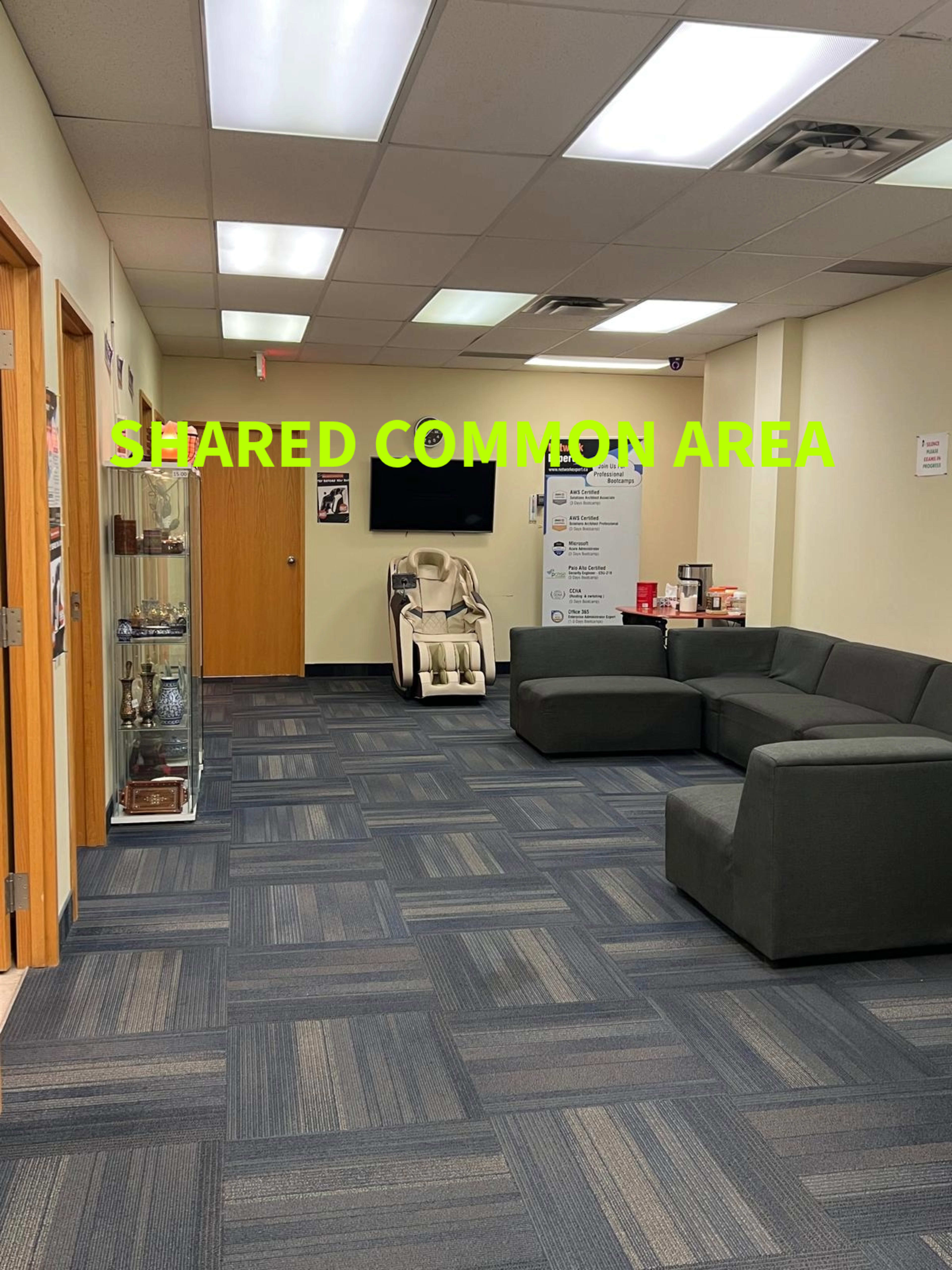 The image shows a spacious shared common area featuring a gray sectional sofa, a relaxation chair, and display cases along with informational posters on the walls.