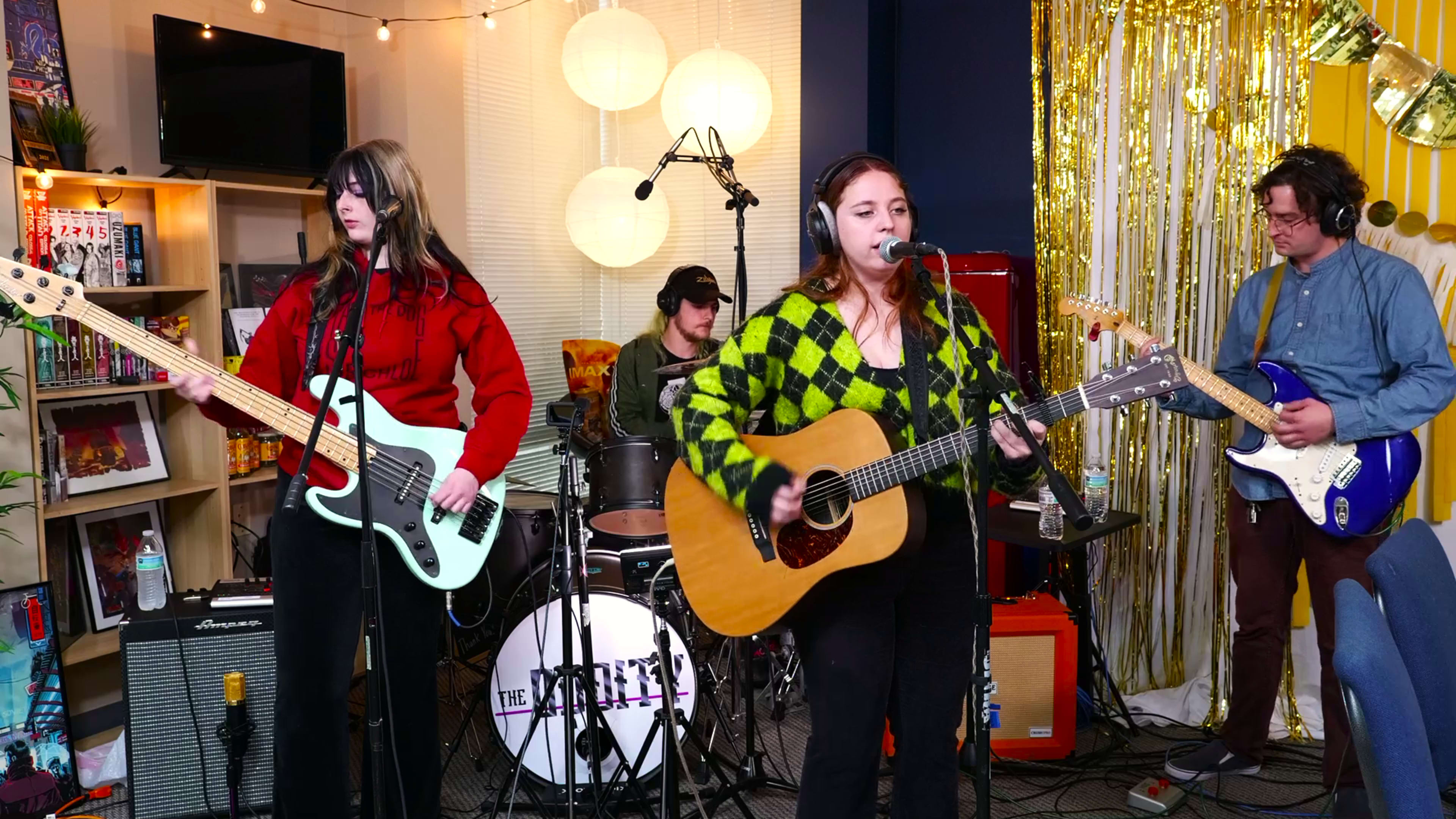A band performs in a decorated space with colorful lighting, featuring a singer with an acoustic guitar, a bassist, and a drummer in the background.