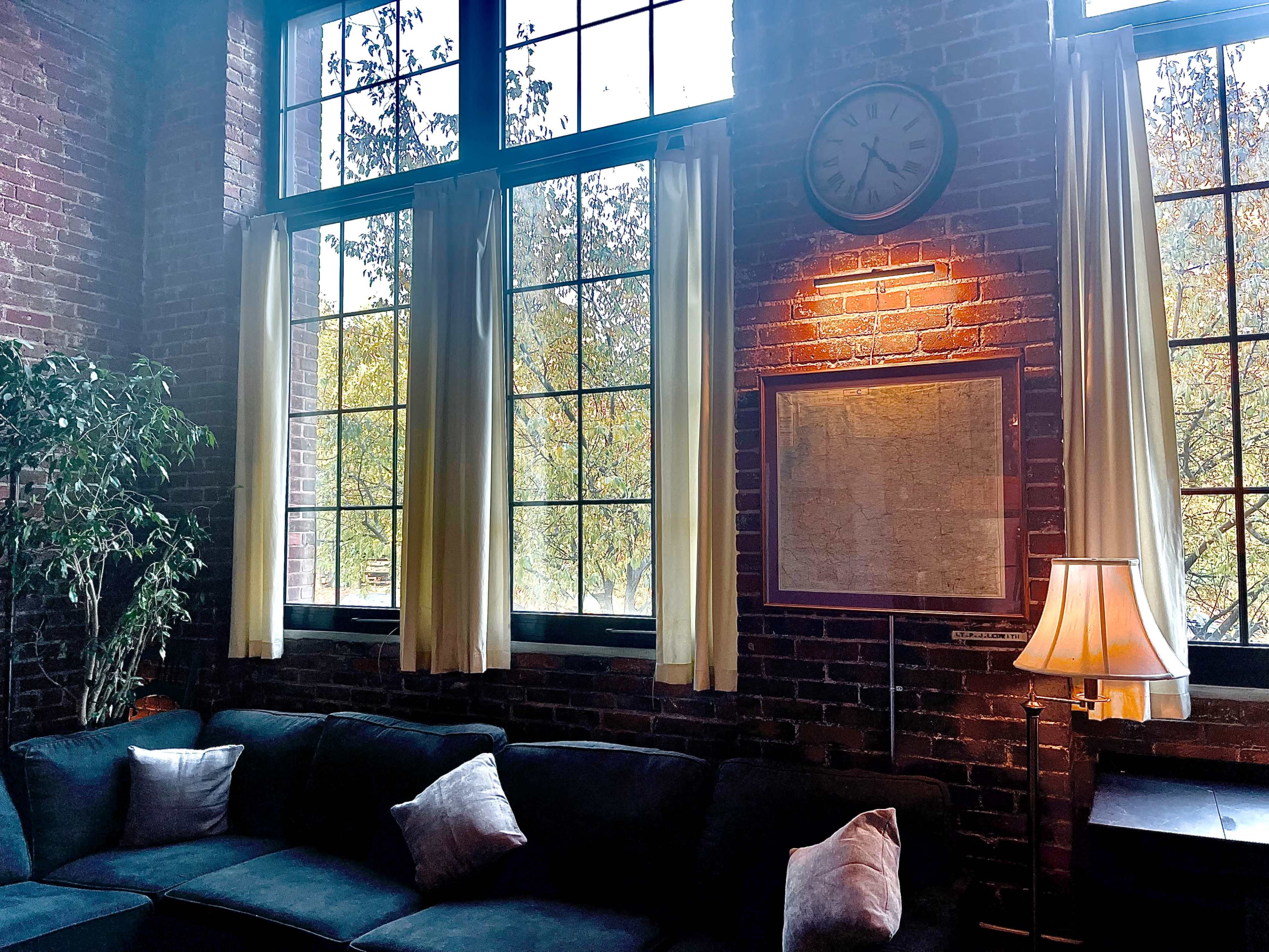 The image shows a cozy living area with a dark sofa, large windows framed by light curtains, and a wall clock above a map.