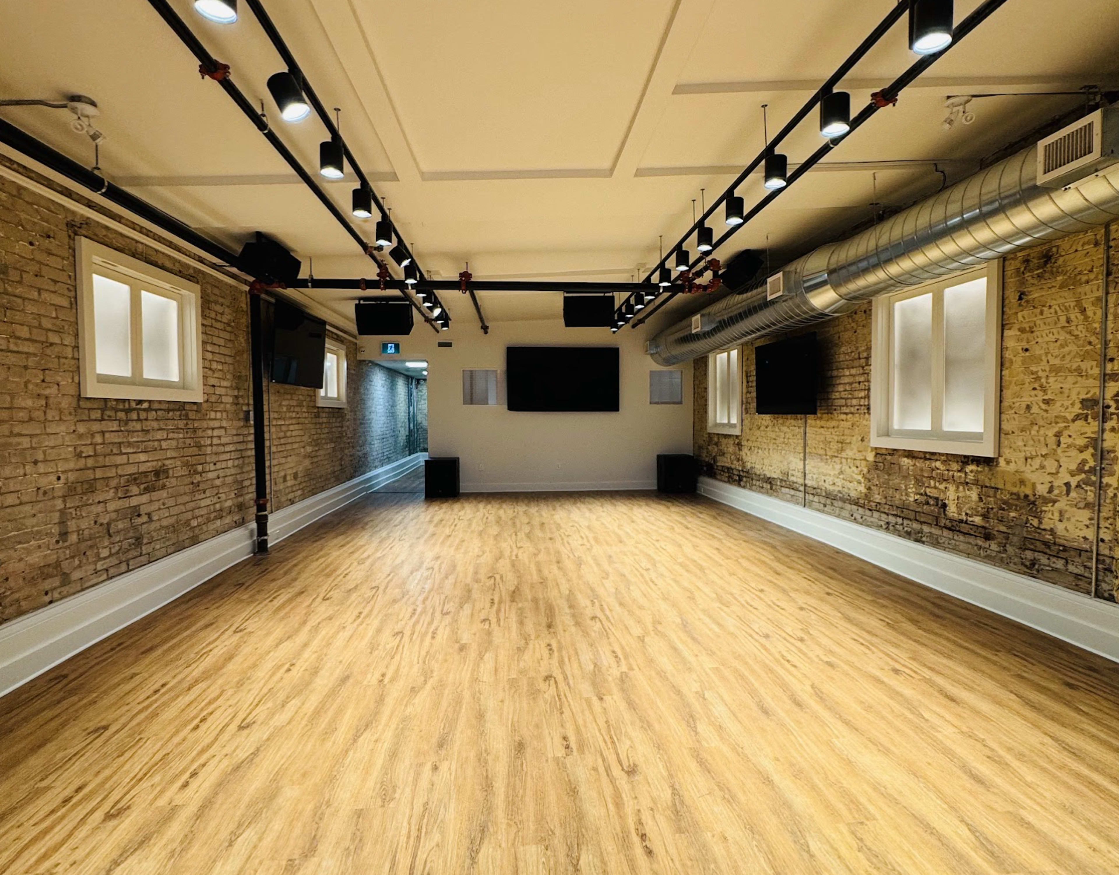 The image shows a spacious room with wooden flooring, exposed brick walls, and several mounted lights and screens.