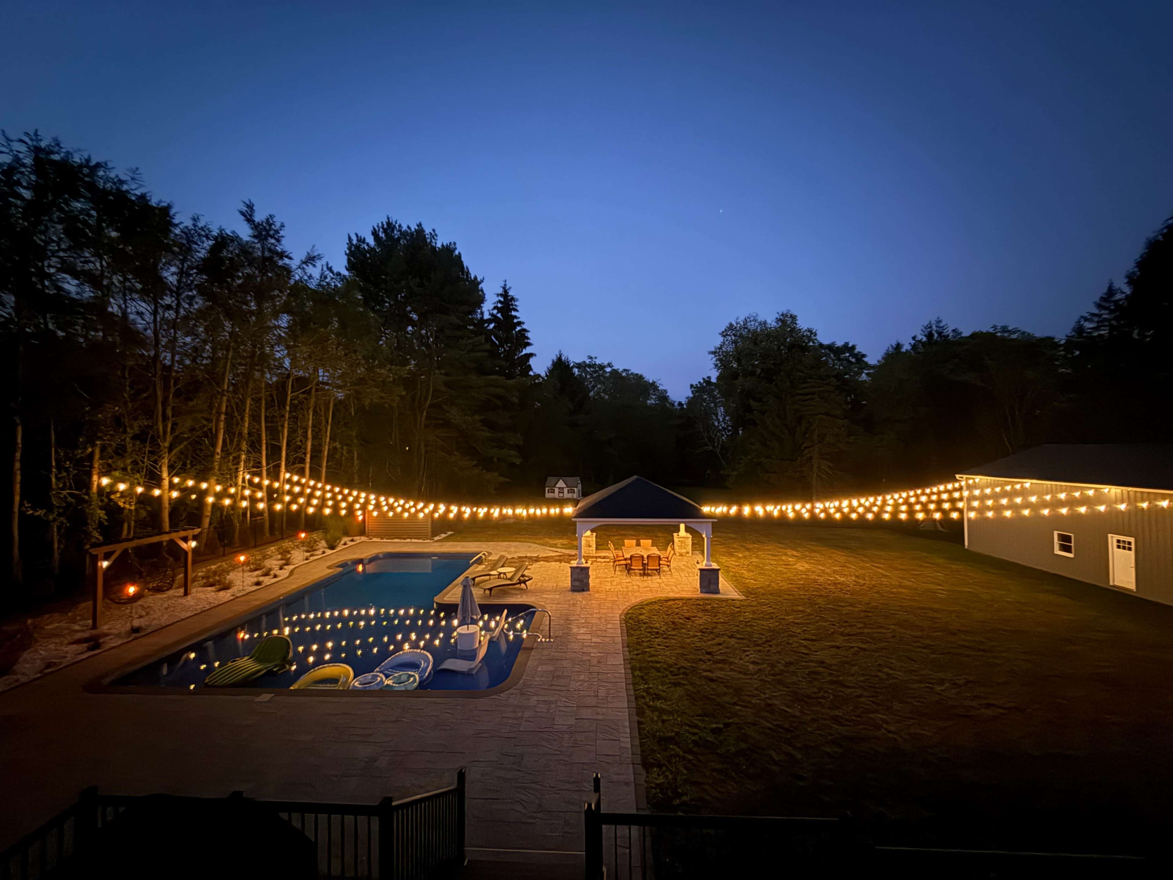 The image shows a backyard scene with a swimming pool, a gazebo lit with string lights, and a grassy area surrounded by trees at dusk.
