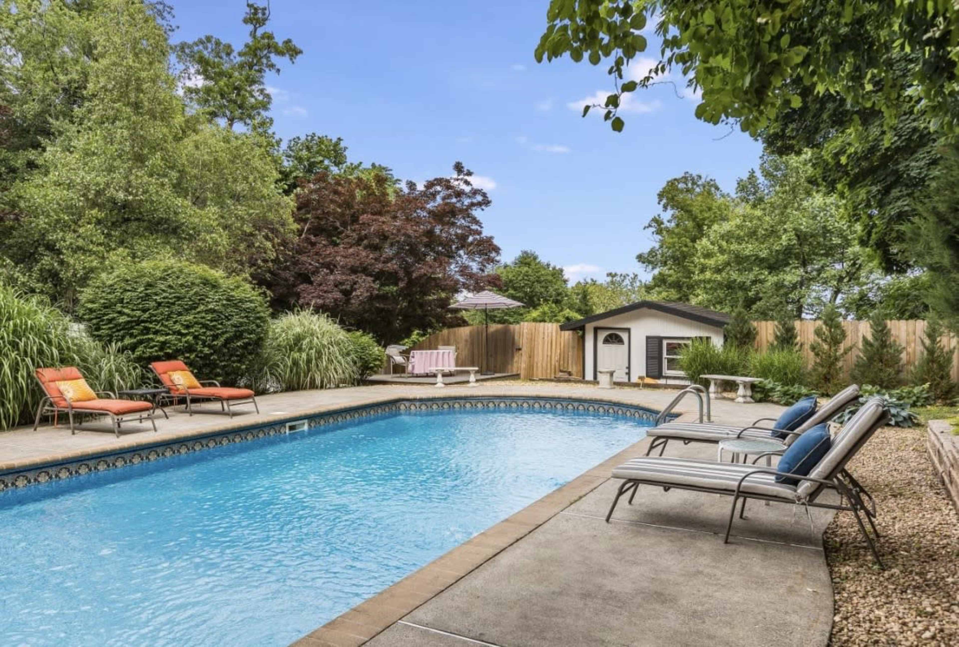 The image shows a backyard pool area surrounded by lounge chairs and greenery, with a small building visible in the background.