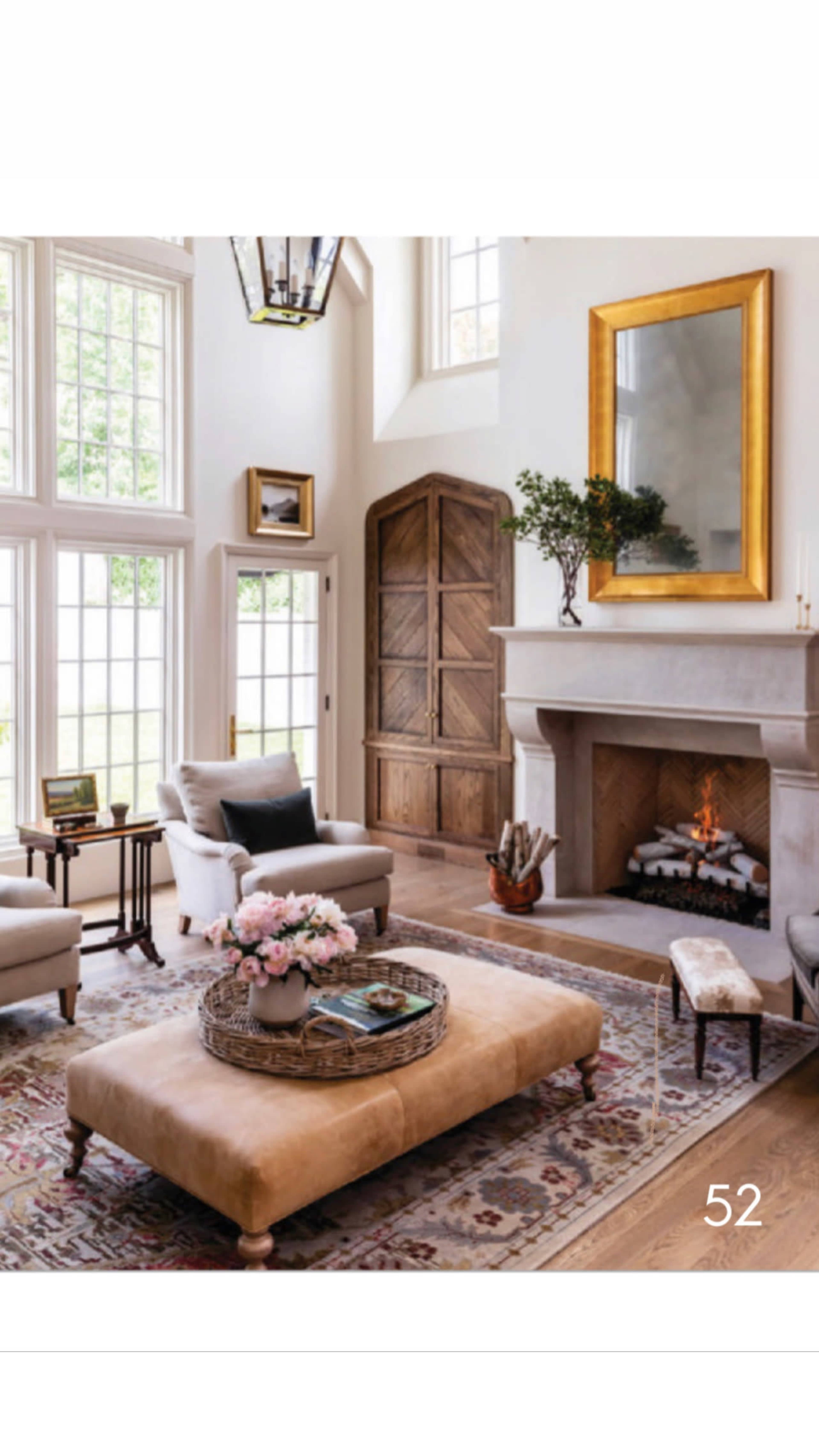 A spacious living room features a large ottoman in the center, surrounded by seating and a fireplace, with natural light streaming through tall windows.