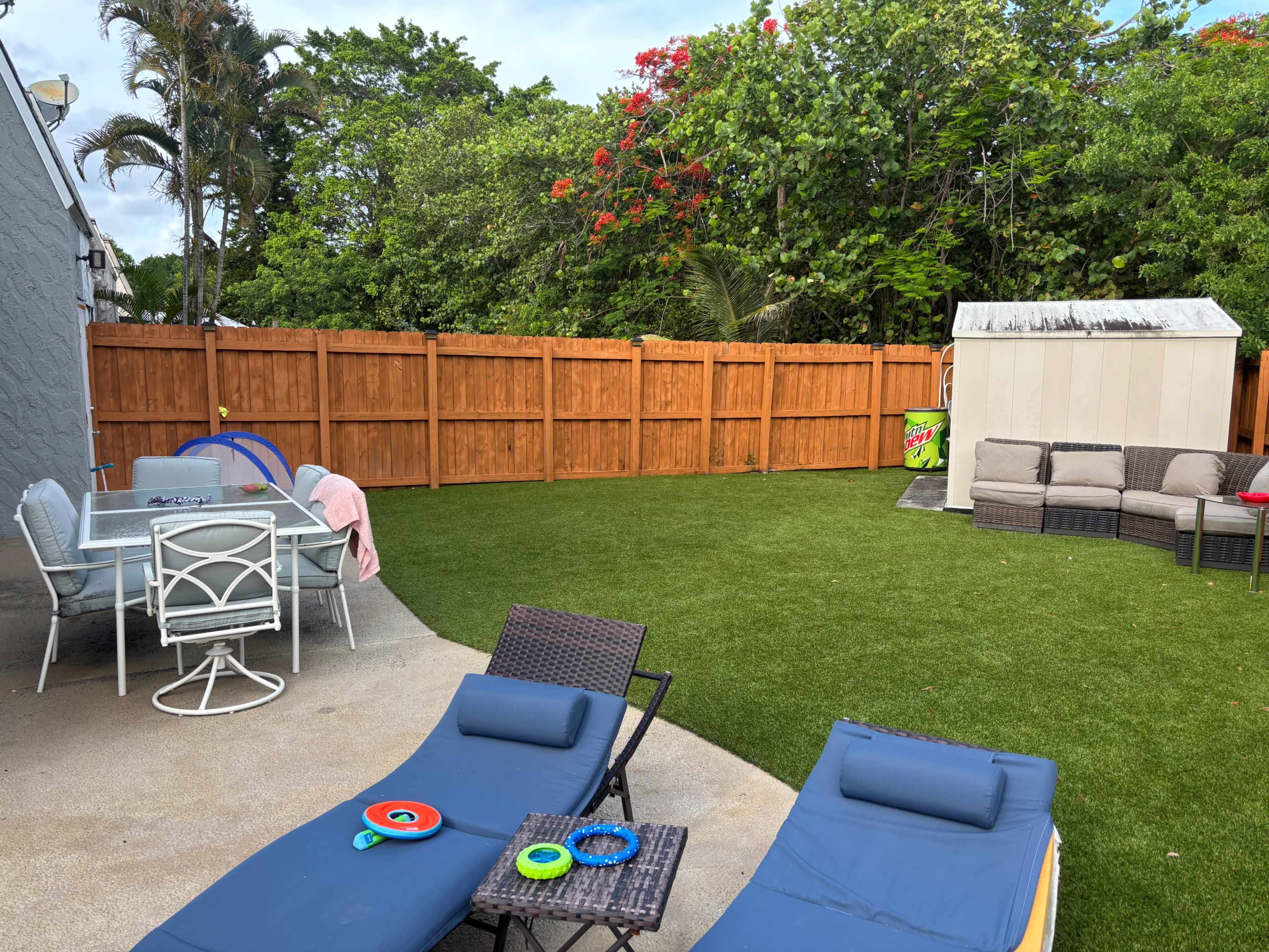 The image shows a backyard with a grassy area, lounge chairs, a dining table, and a storage shed surrounded by a wooden fence.
