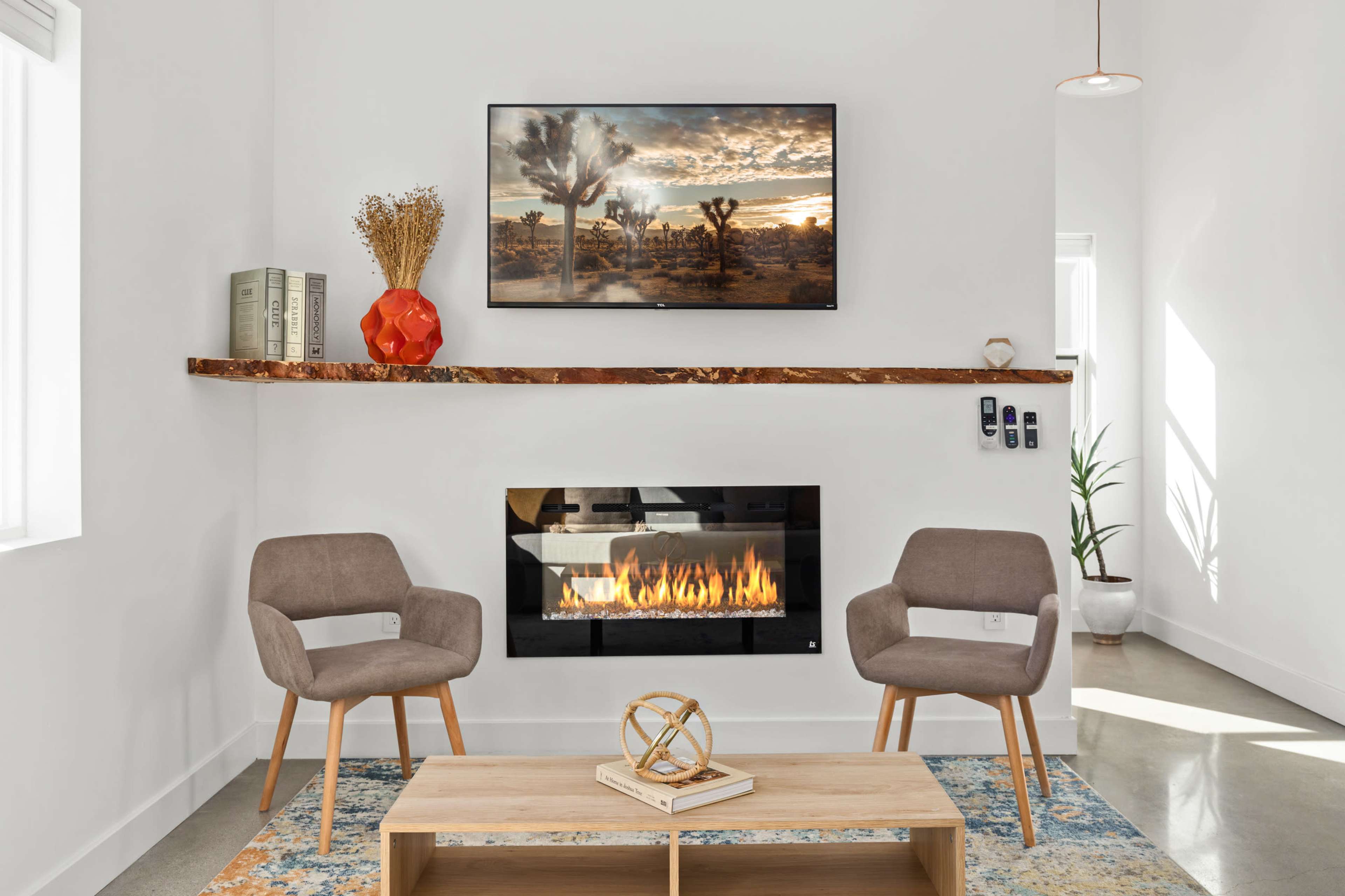 The image shows a modern living room with two gray chairs, a coffee table, a wall-mounted television displaying a landscape, and a fireplace beneath it.