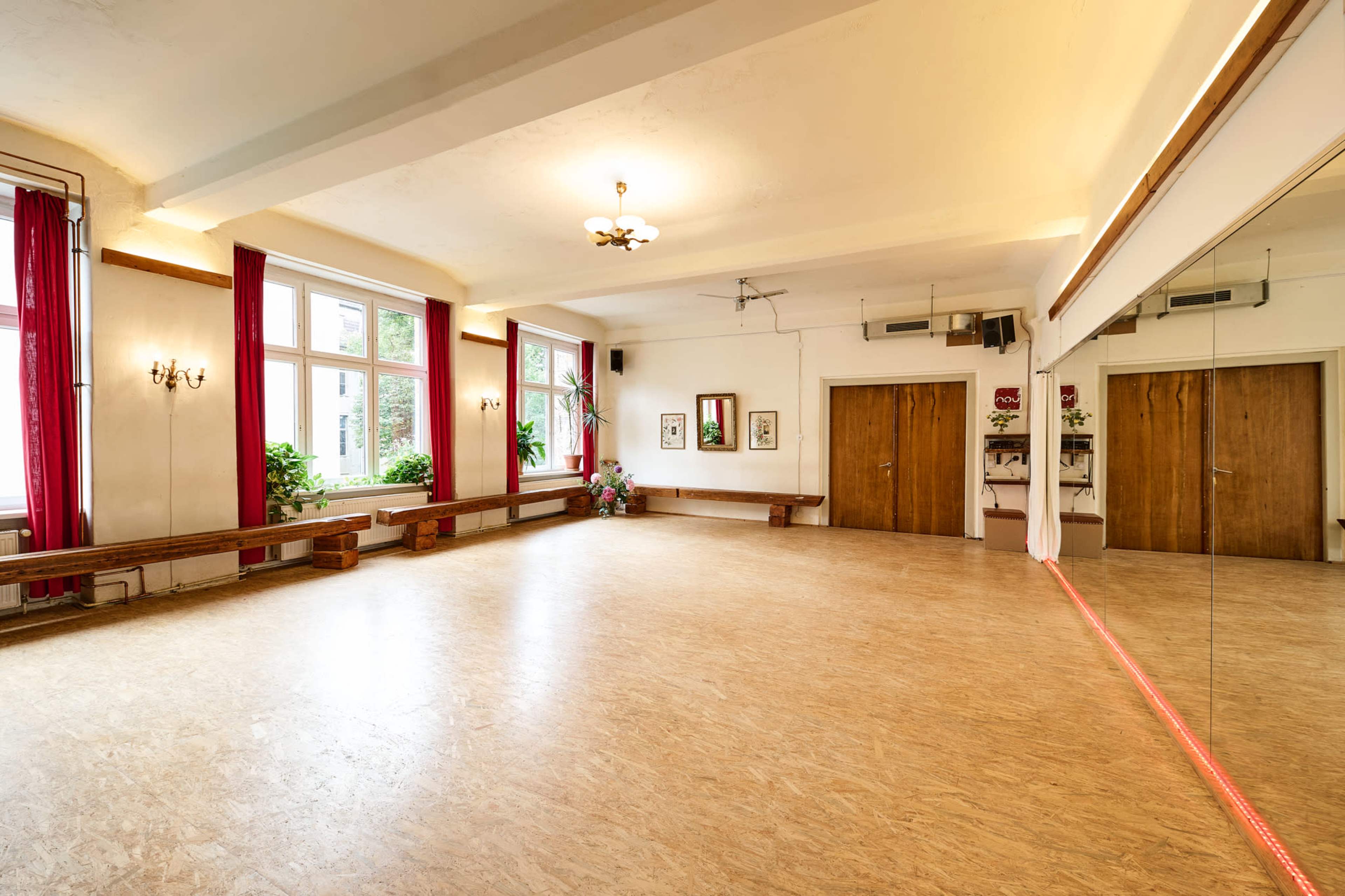 The image shows a spacious, well-lit room with wooden floors, large windows, and a mirror along one wall, featuring simple furnishings and decorative plants.