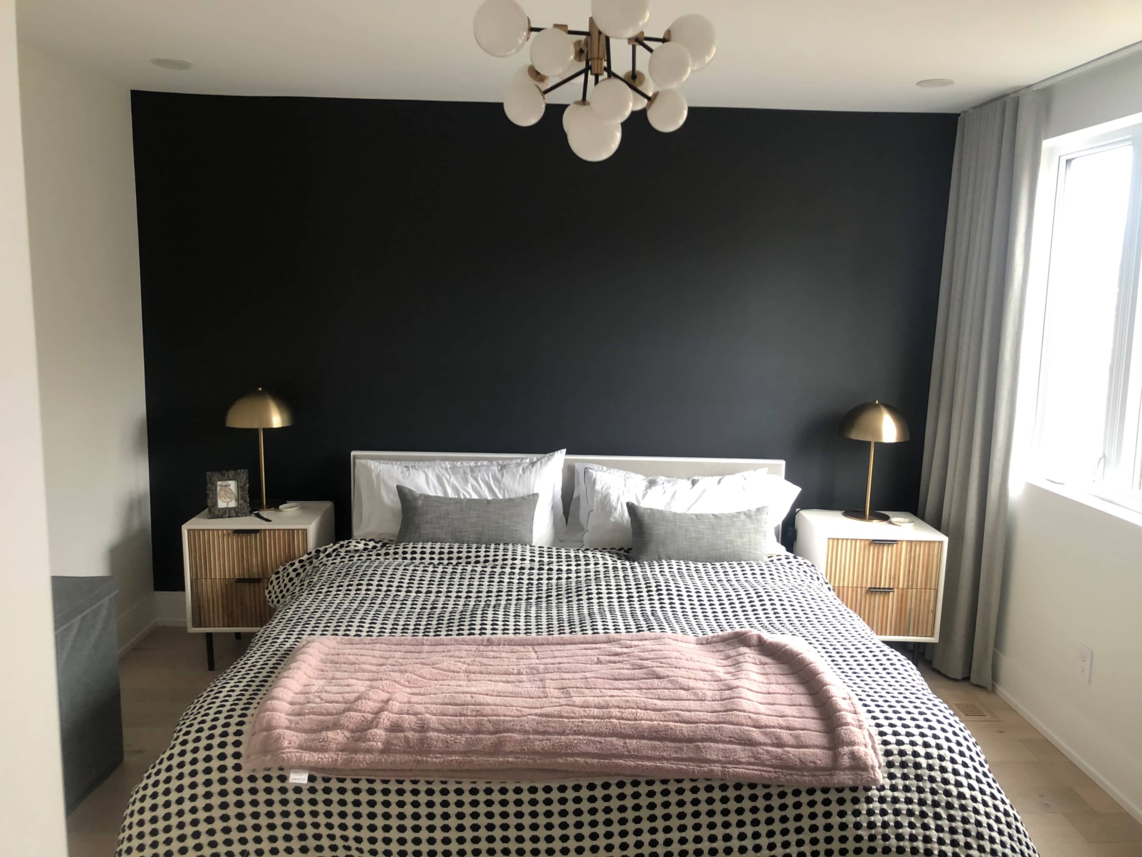 The image shows a modern bedroom featuring a bed with a black and white polka dot comforter, two gold bedside lamps, and a large window with light-colored curtains.