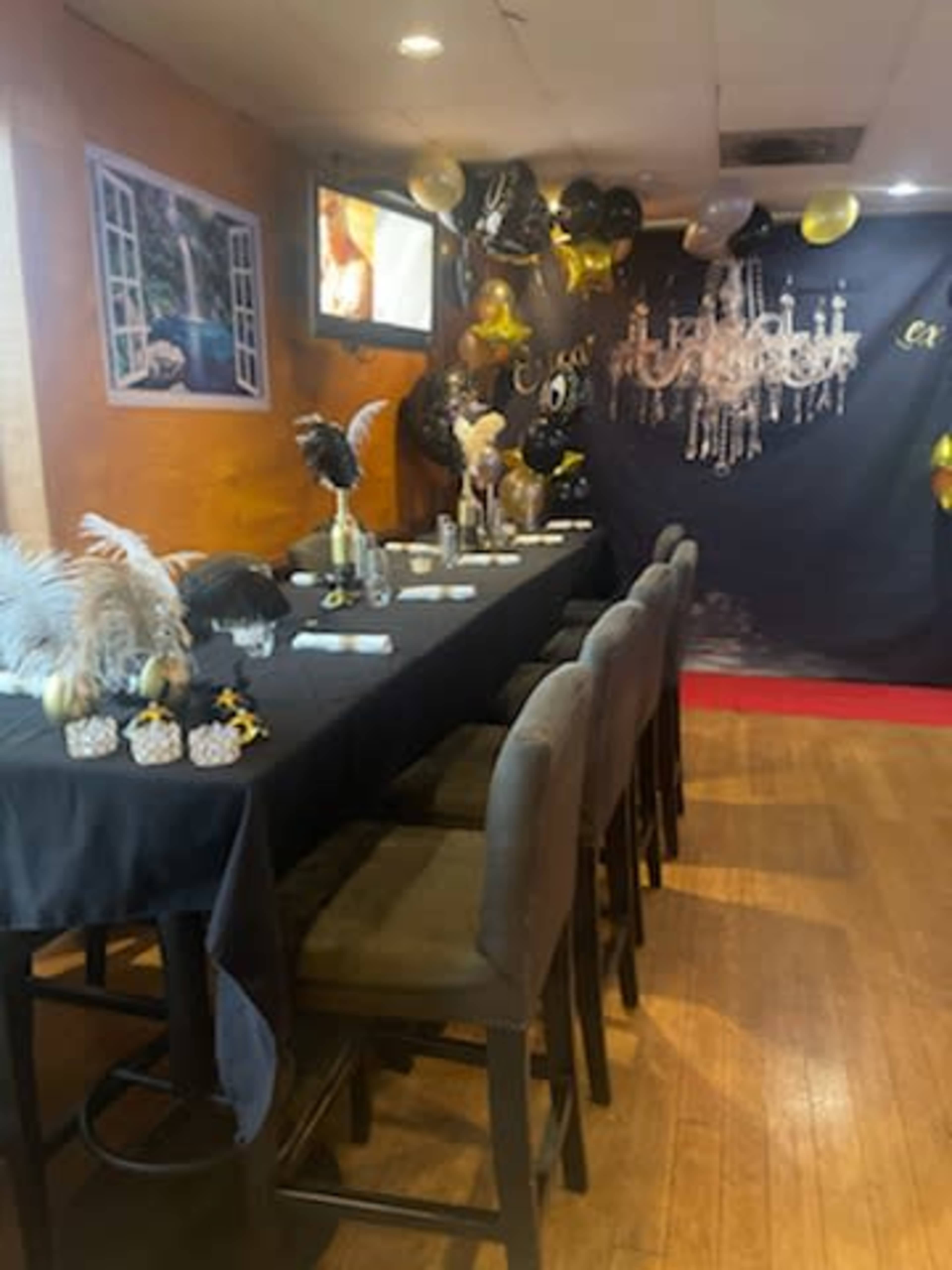 A dining area decorated with black and gold balloons, a long dark table set with utensils, and a background featuring a chandelier and a television screen.