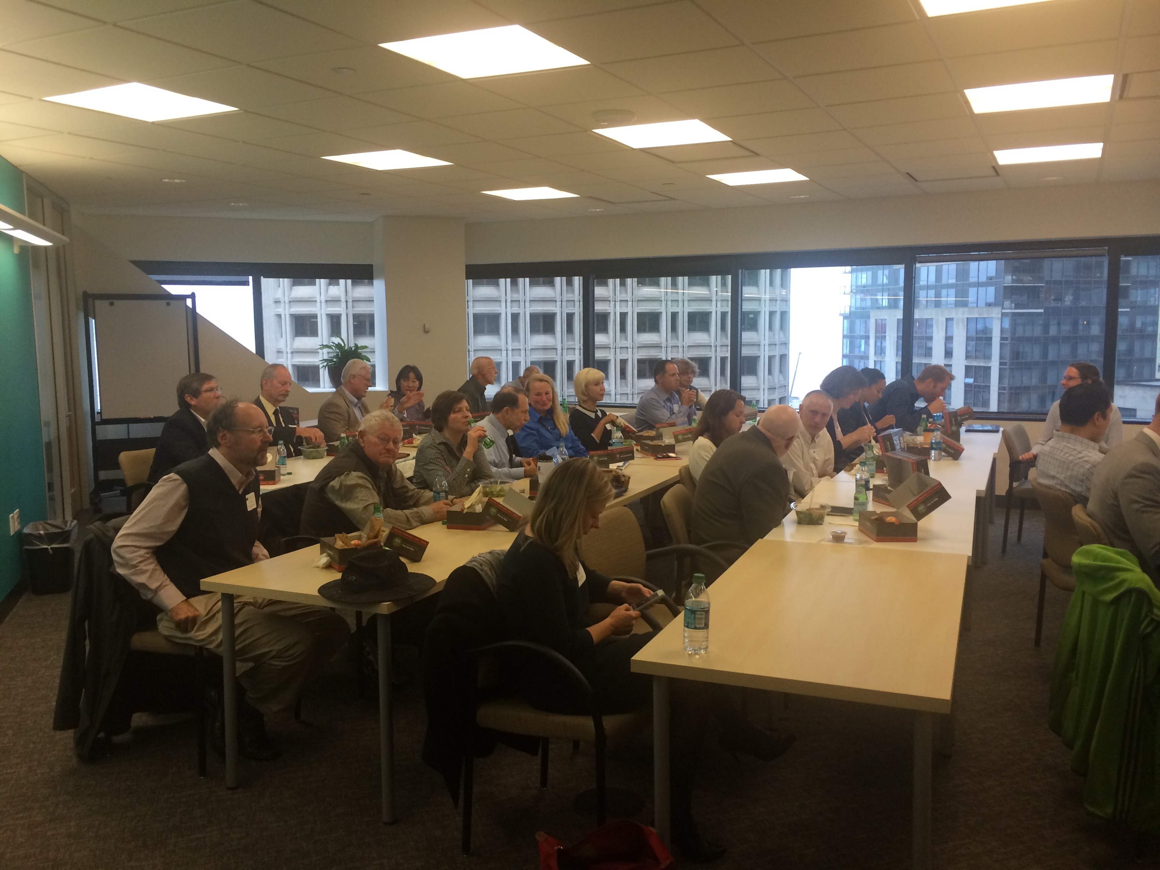 A group of people are seated around tables in a conference room, some looking at electronic devices, while others are engaged in discussion.